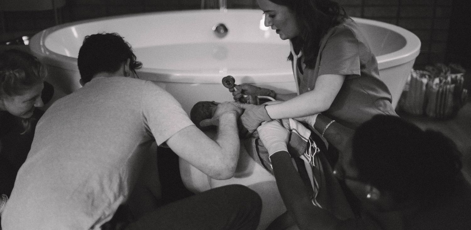 Black and white photo of birth team assisting woman during water birth in tub.