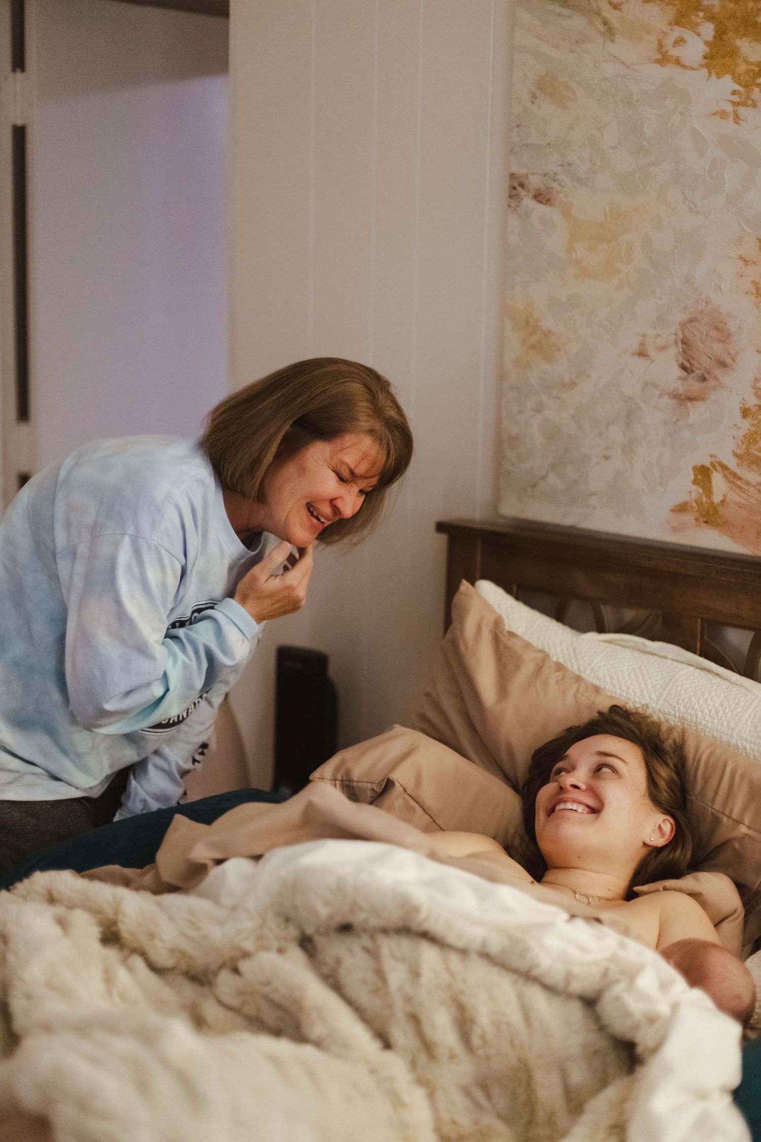 A mother laughs while waking her smiling teenage daughter in a cozy bedroom.