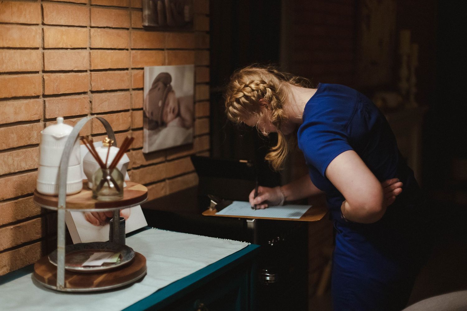 Woman in blue dress signing documents at a teal desk beside candles and art supplies in a warmly lit room.