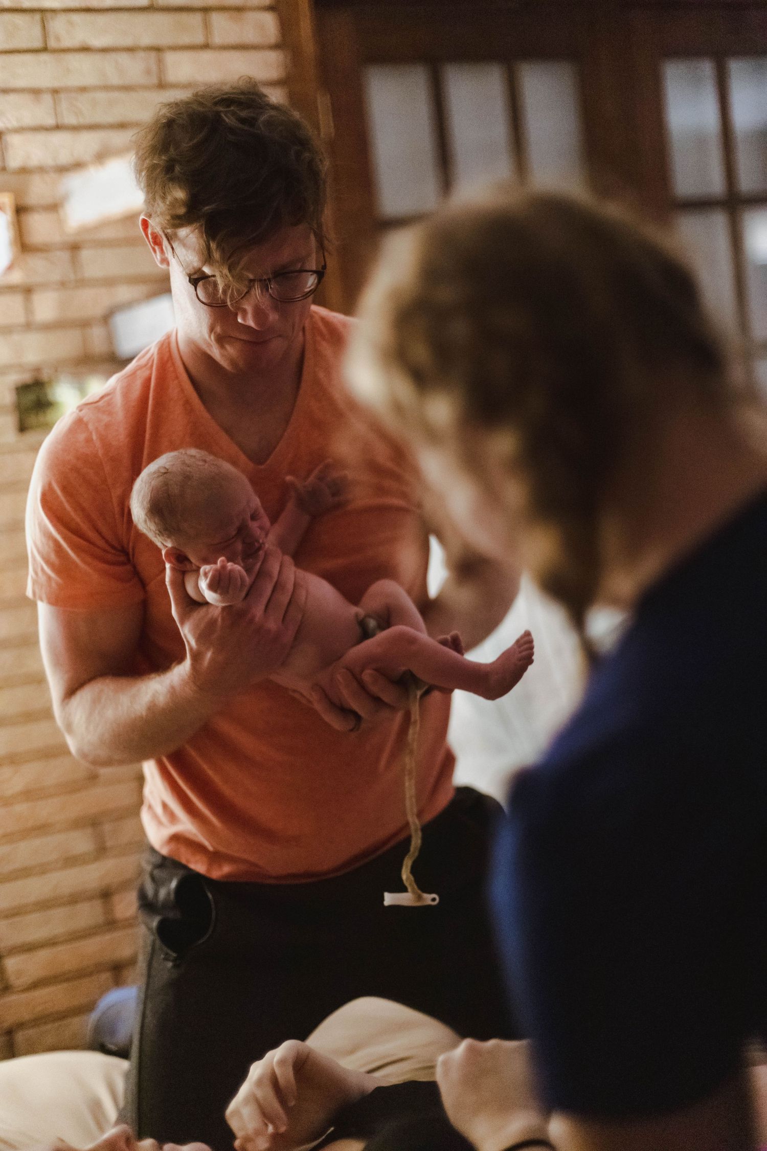 A father in an orange shirt holds his newborn baby with umbilical cord still attached during home birth.