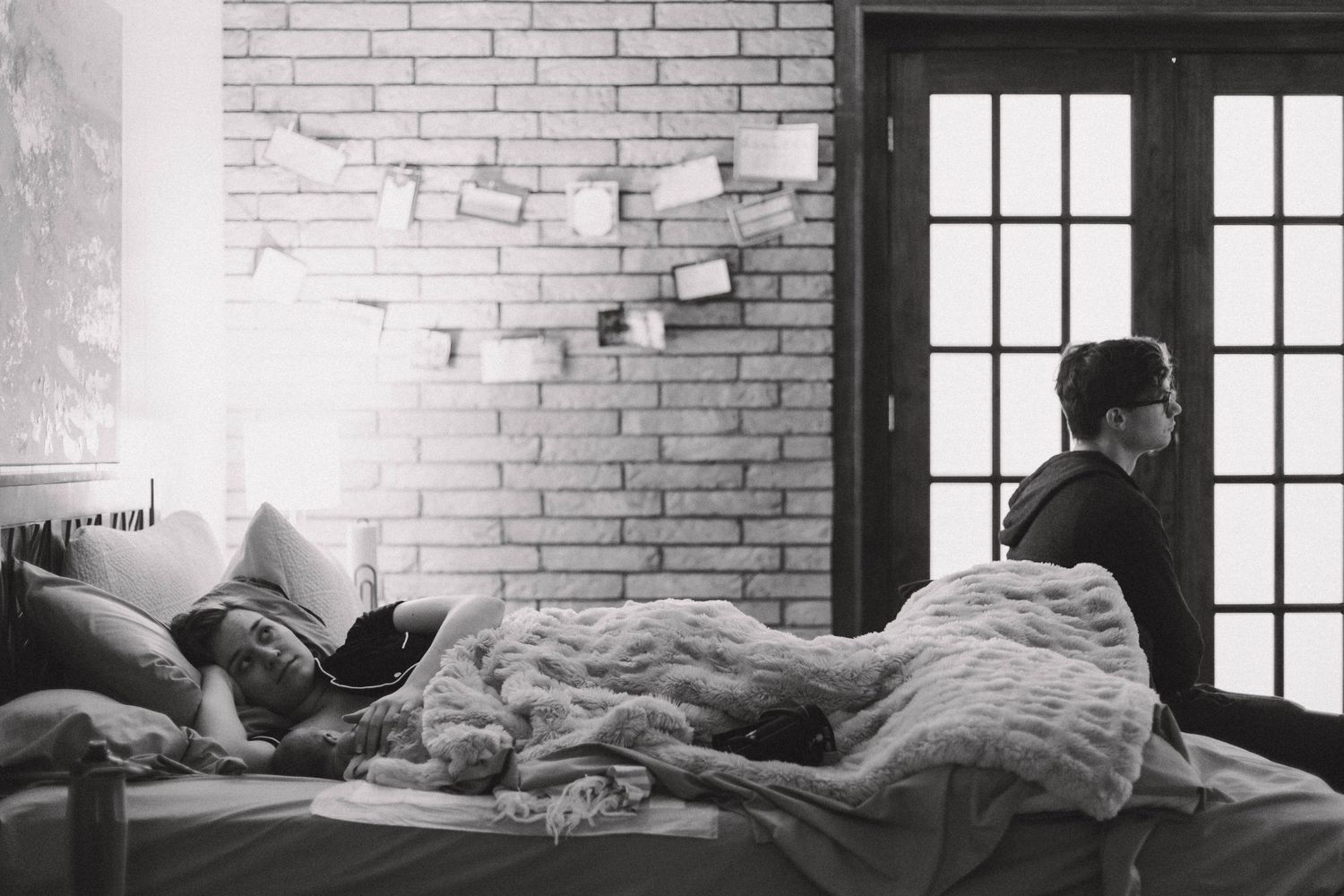Black and white photo of a person sleeping in bed while another stands by a window.