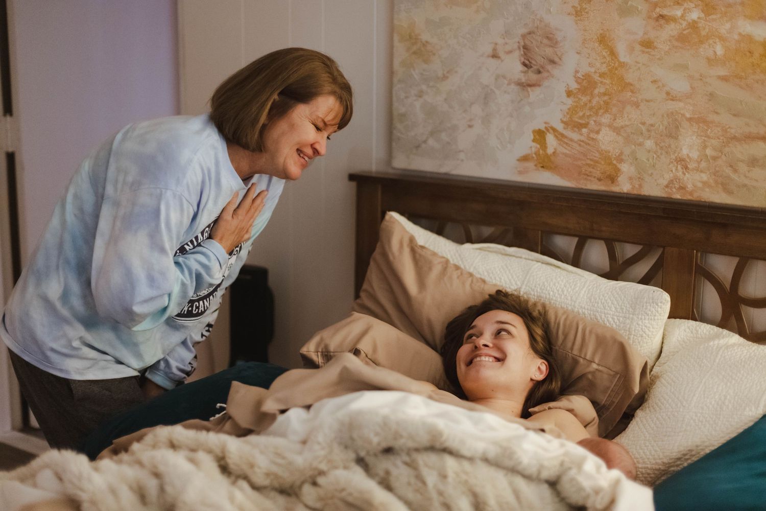 A caring older woman leans over to speak with a smiling young mother resting in bed after childbirth.