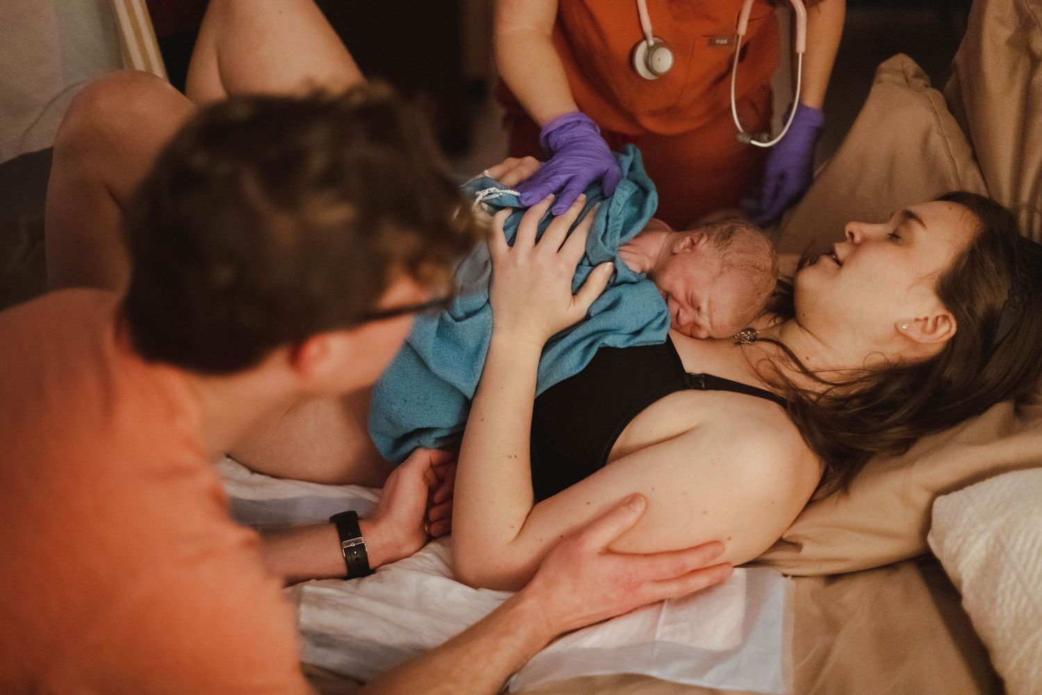 A mother receives her newborn baby while lying in bed, surrounded by a supportive partner and medical team.