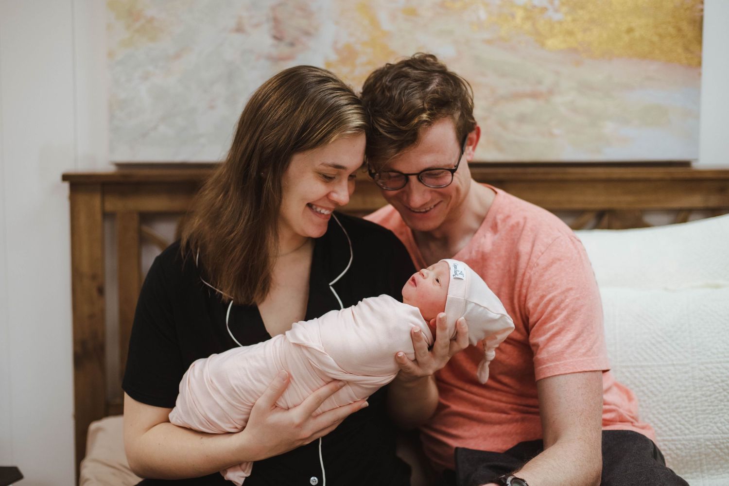 Happy new parents smile while holding their swaddled newborn baby together on a bed.