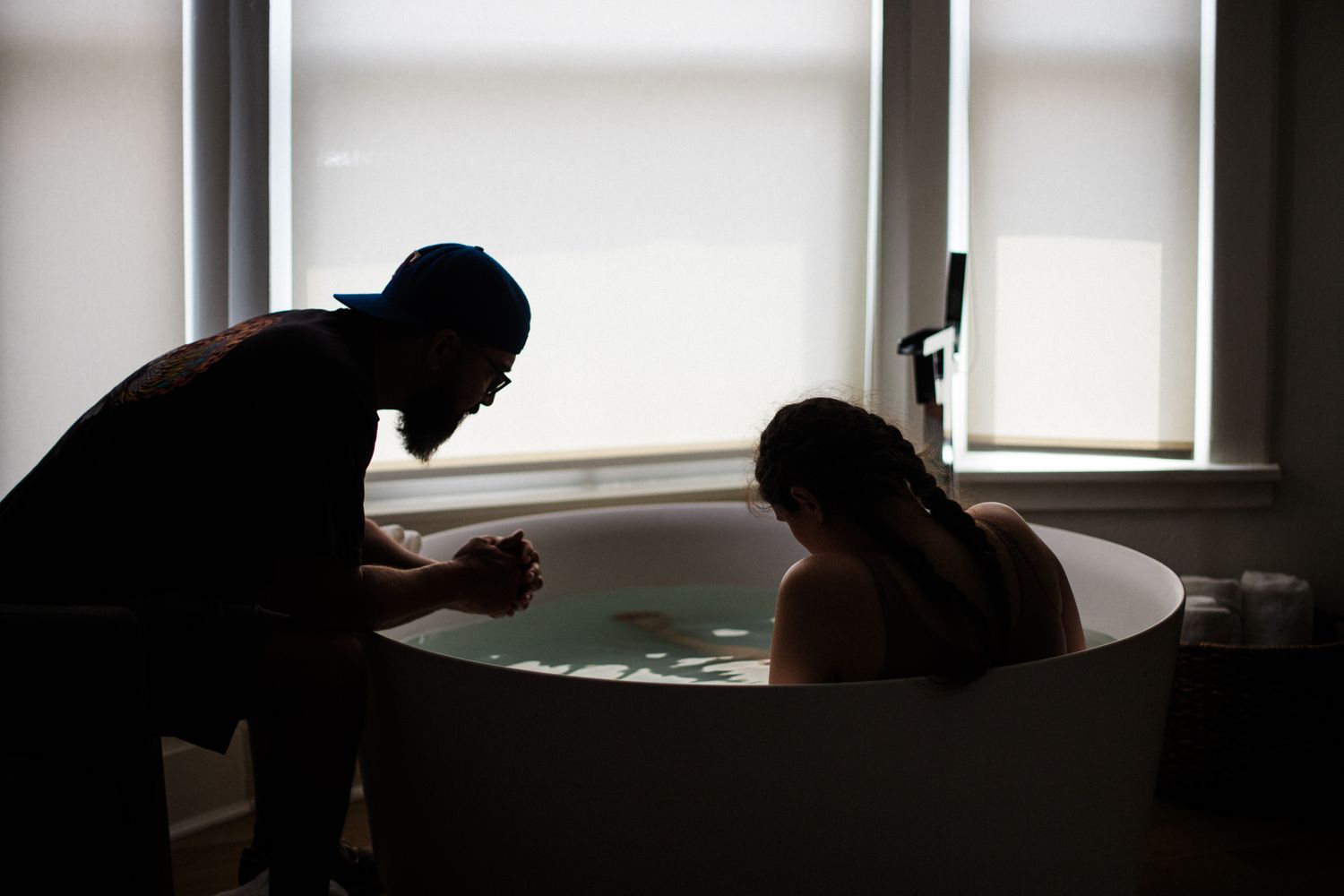 Silhouette of a support person kneeling beside a woman in a birthing tub during labor.