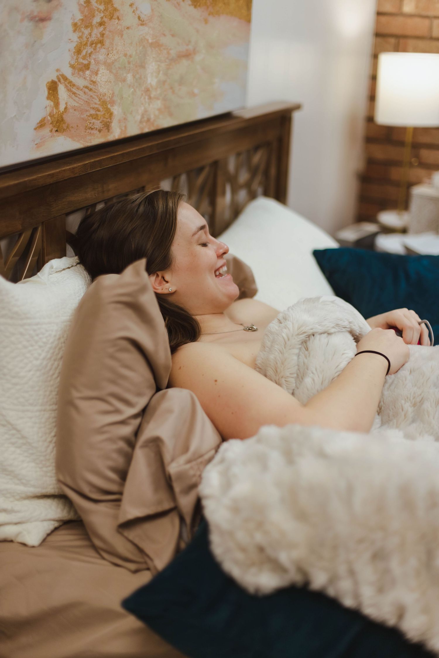 A smiling new mother rests comfortably in bed surrounded by soft pillows after giving birth at home.