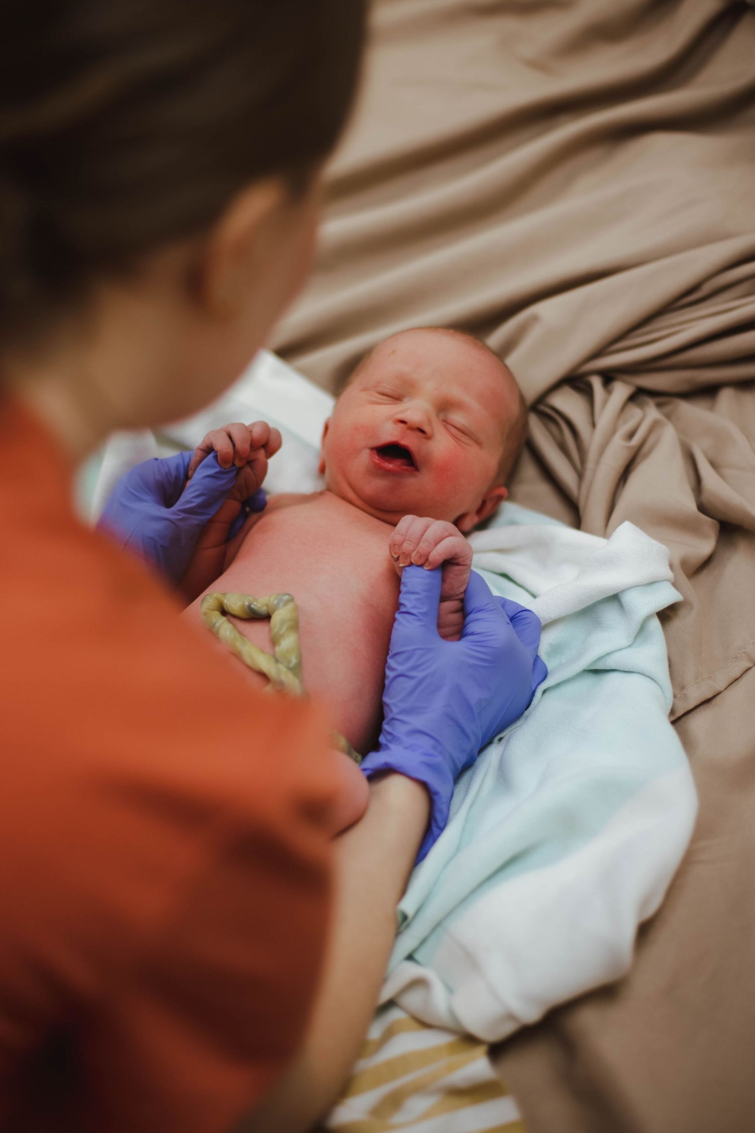 Midwife in orange scrubs and purple gloves examining alert newborn baby after home birth delivery.