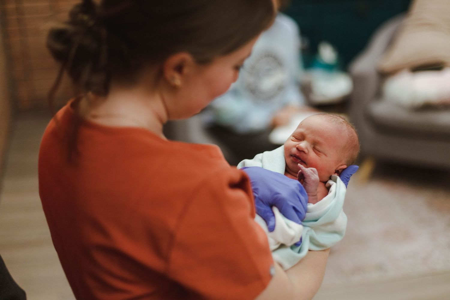 A midwife in orange scrubs gently holds and presents a smiling newborn baby after a home delivery.