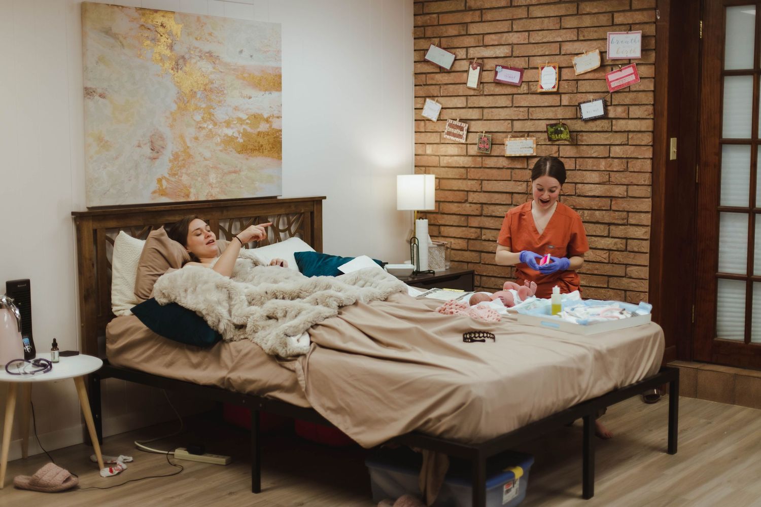 A midwife in orange scrubs tends to a newborn while the mother rests in bed after a home birth.