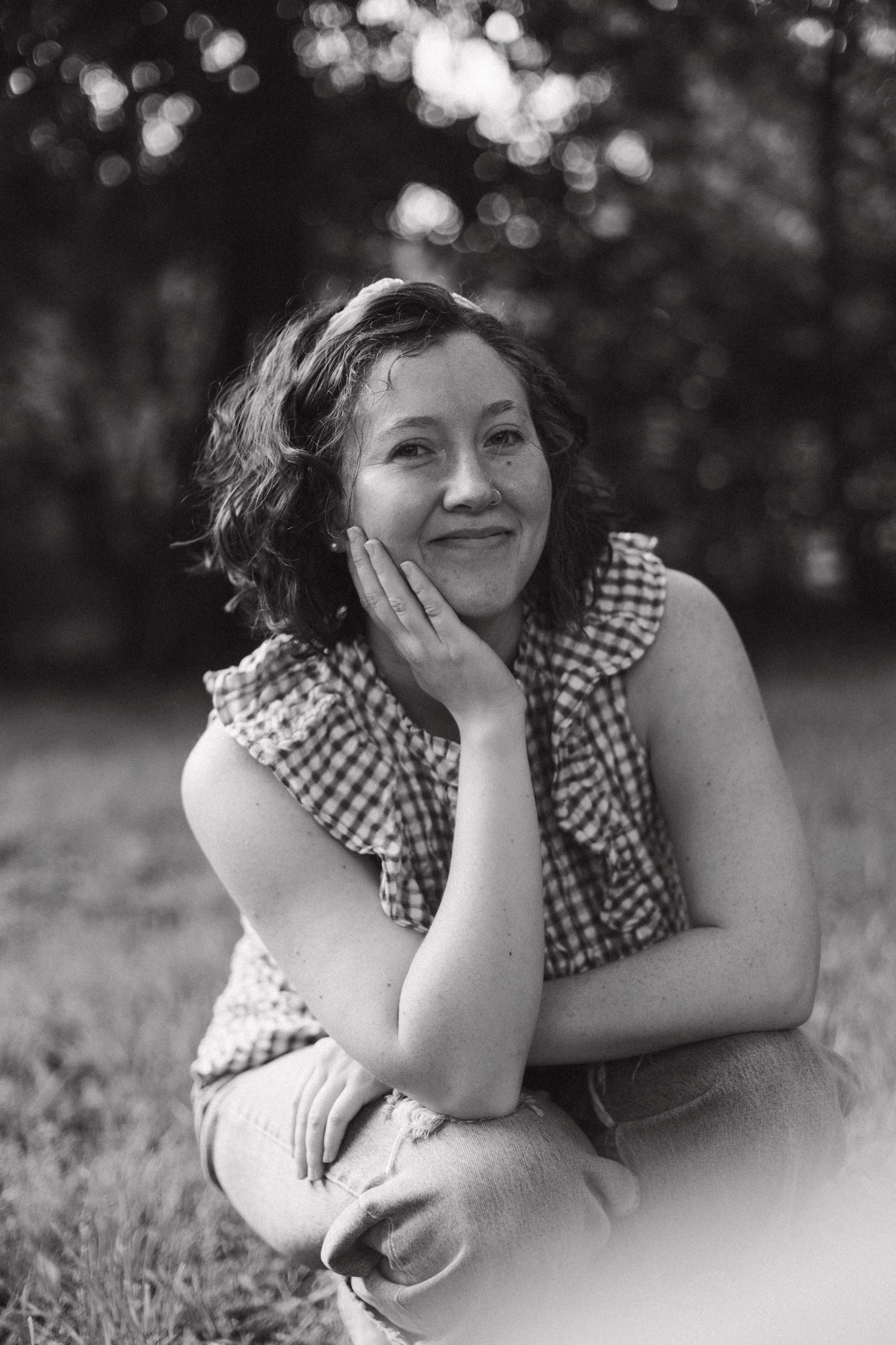 Black and white portrait of a smiling woman sitting outdoors in a gingham top.
