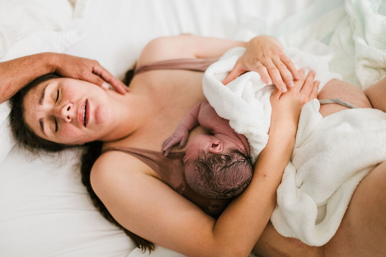 New mother resting on white bed sheets while embracing her newborn baby after delivery.