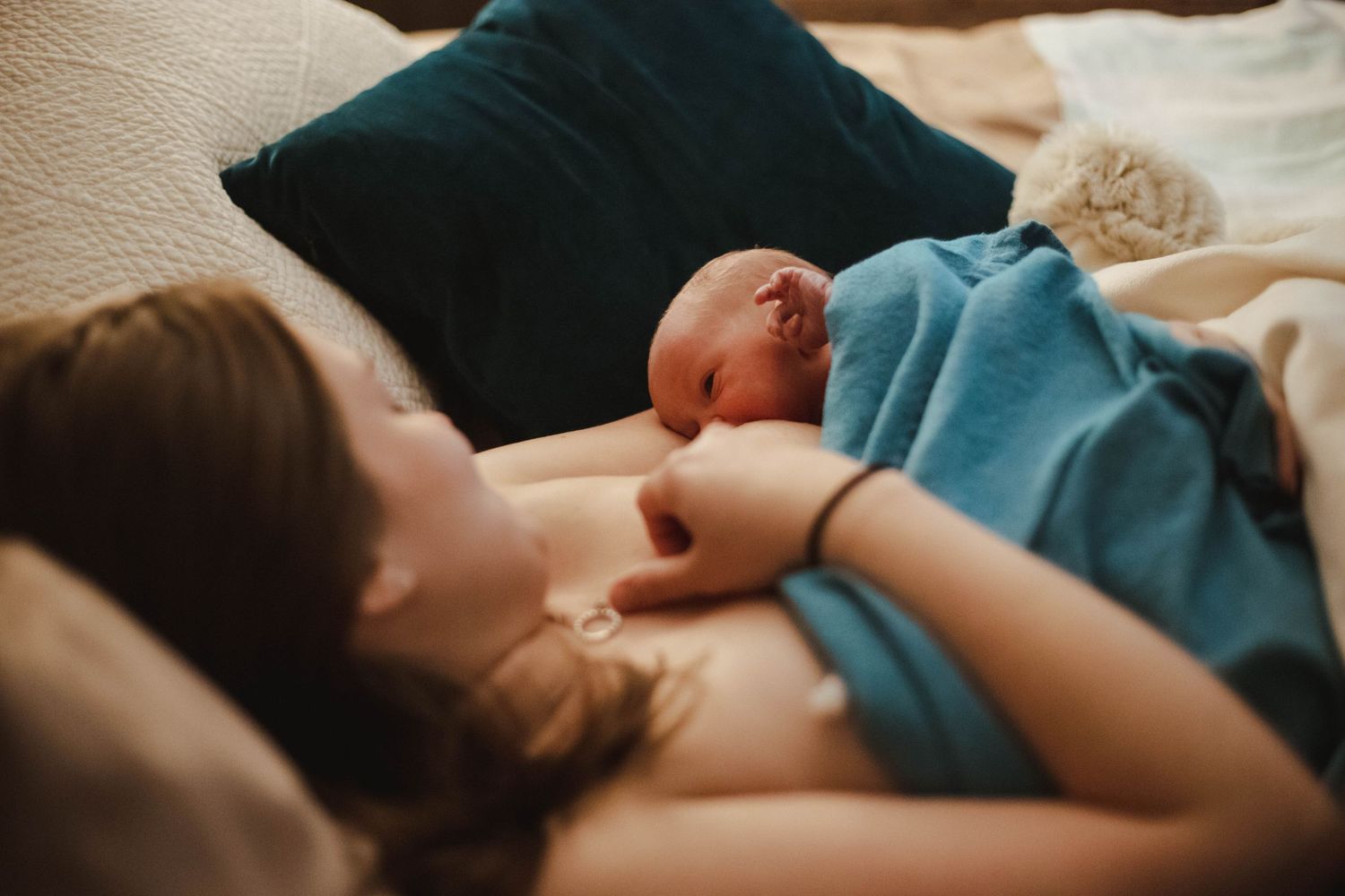 Mother resting in bed while breastfeeding her newborn baby wrapped in a teal blanket after home birth.