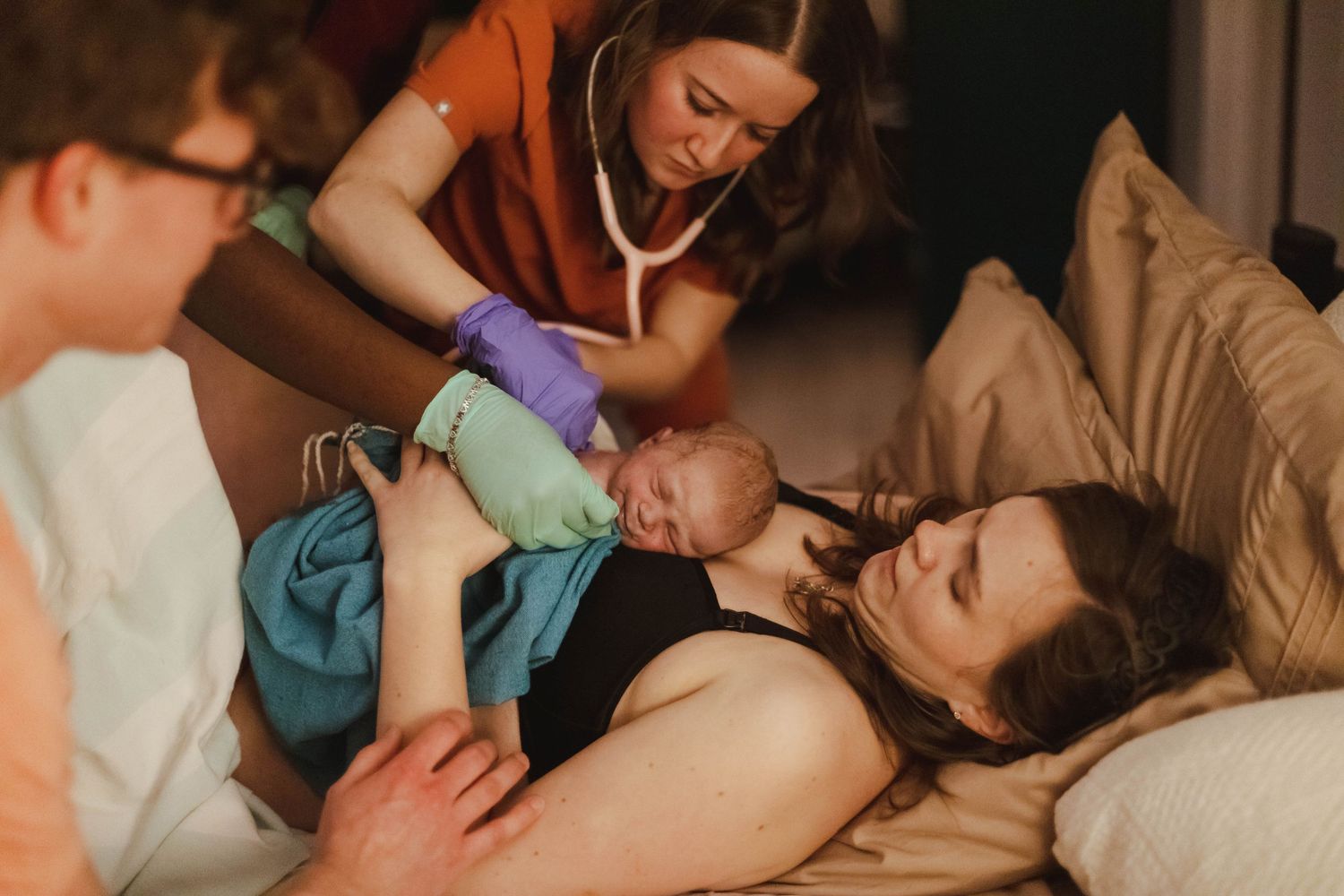 A midwife checks a newborn with a stethoscope as a mother cradles her baby after home birth.