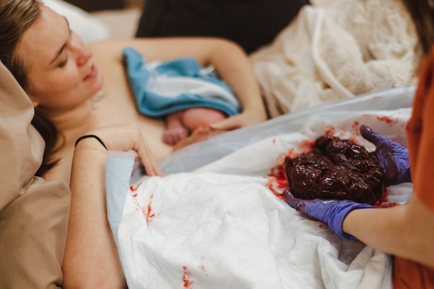 Mother holding newborn baby after childbirth with medical staff delivering placenta in foreground.