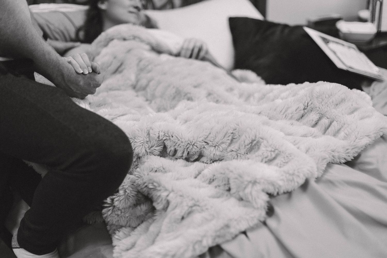 Black and white close-up of partner holding laboring mothers hand during home birth on fluffy blanket.