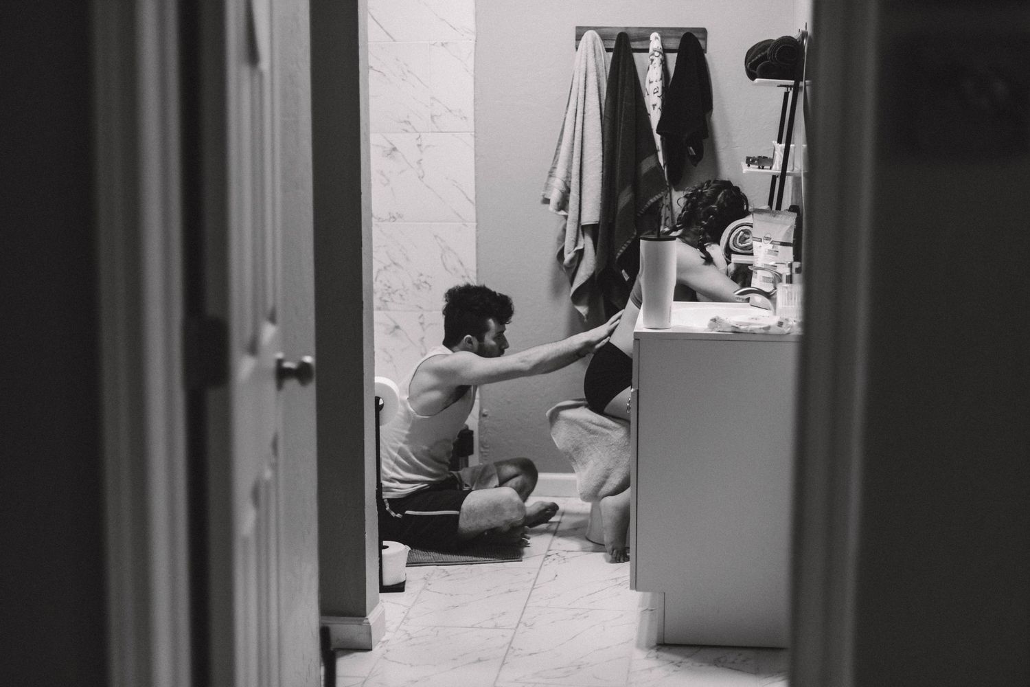 Birth partner comforting laboring woman on bathroom floor, candid black and white image.