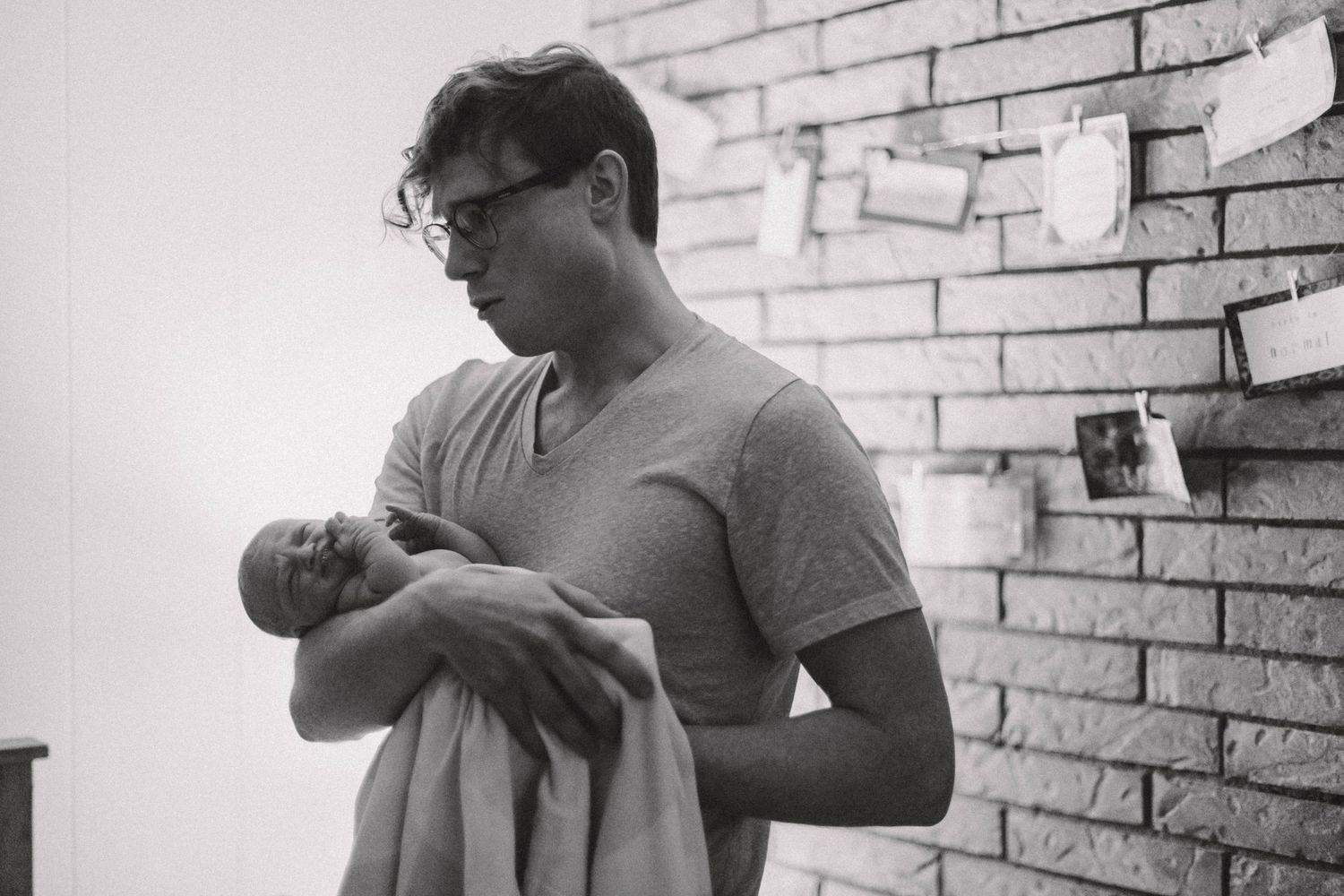 Black and white photo of a new father wearing glasses, cradling his newborn baby against a brick wall.
