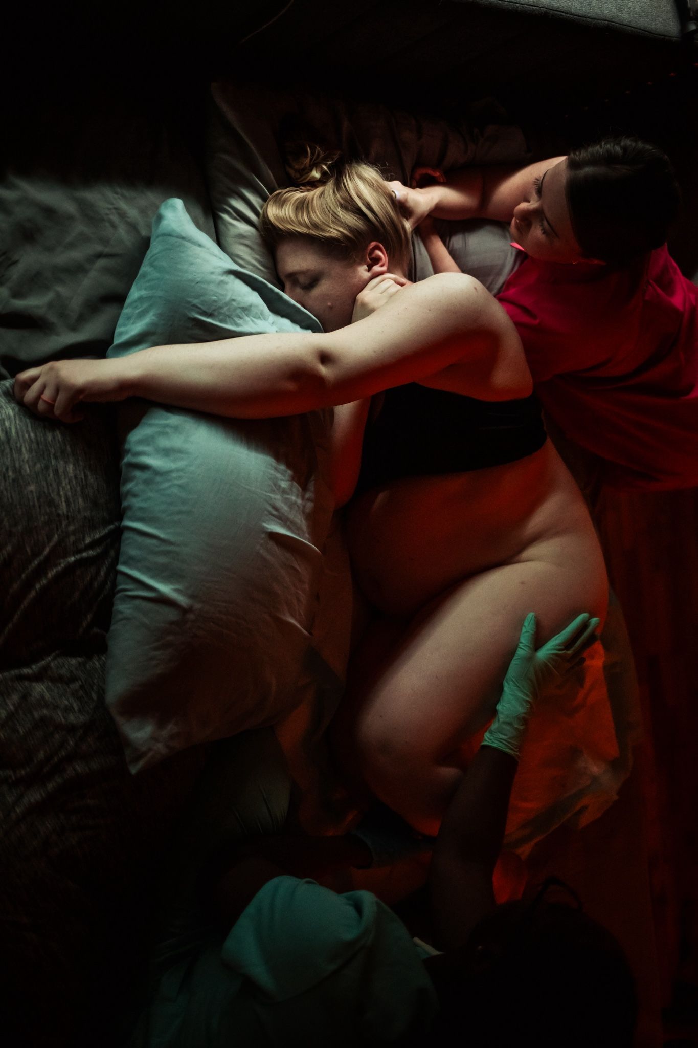 Pregnant woman sleeping on her side in bed, cradled by a partner in a dimly lit bedroom.