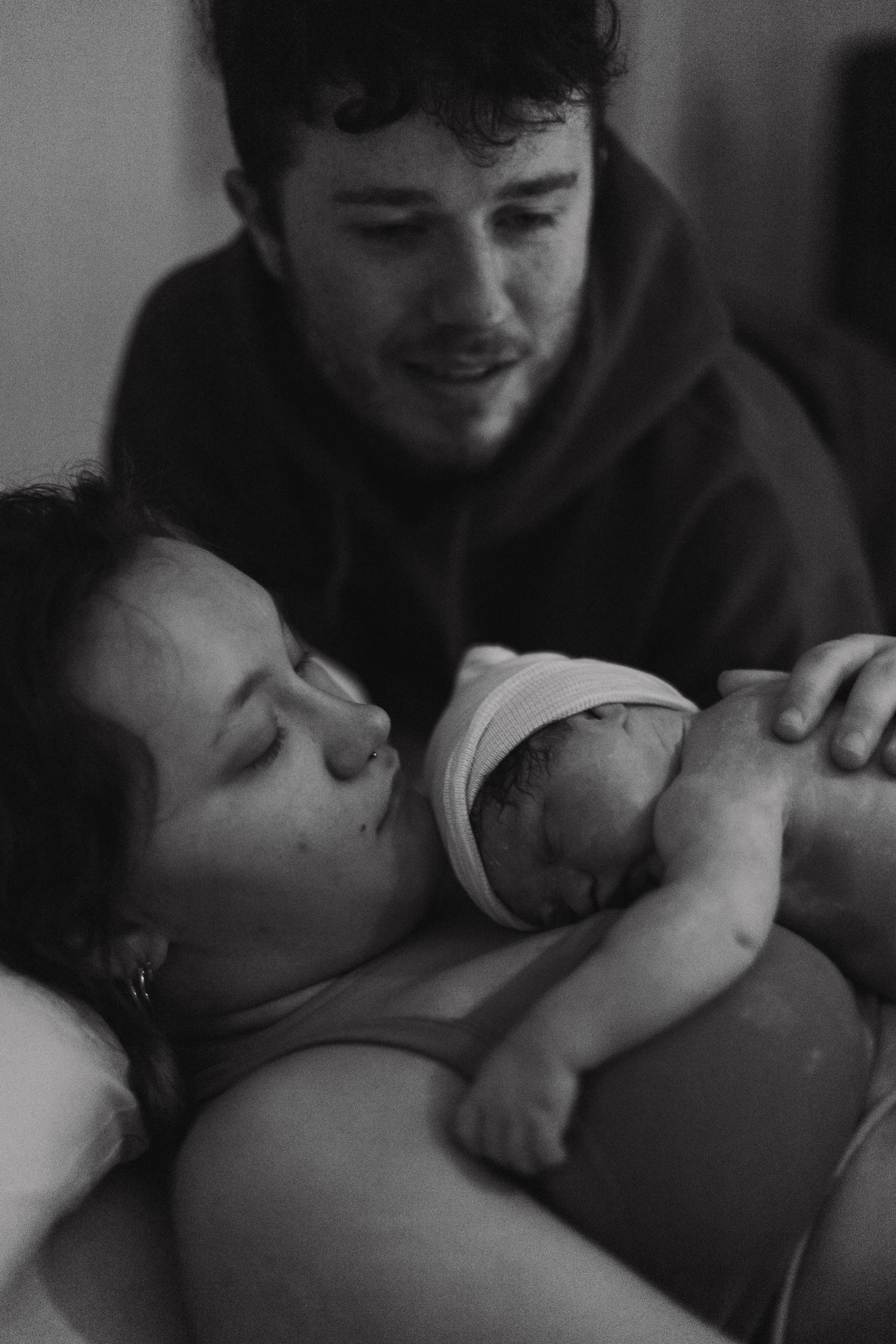 Black and white photo of new parents holding their swaddled newborn baby in a hospital room.