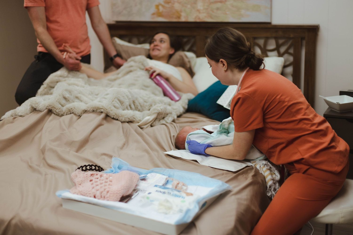 Midwife in orange scrubs attending to new mother resting in bed after home birth with partner nearby.