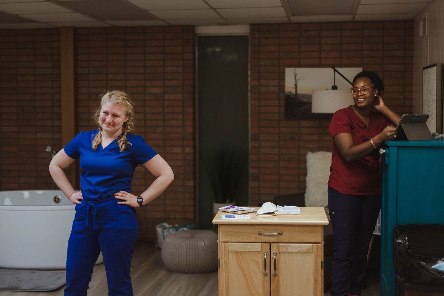 Two midwives smile and relax in a birth center room equipped with a birthing tub and medical supplies.