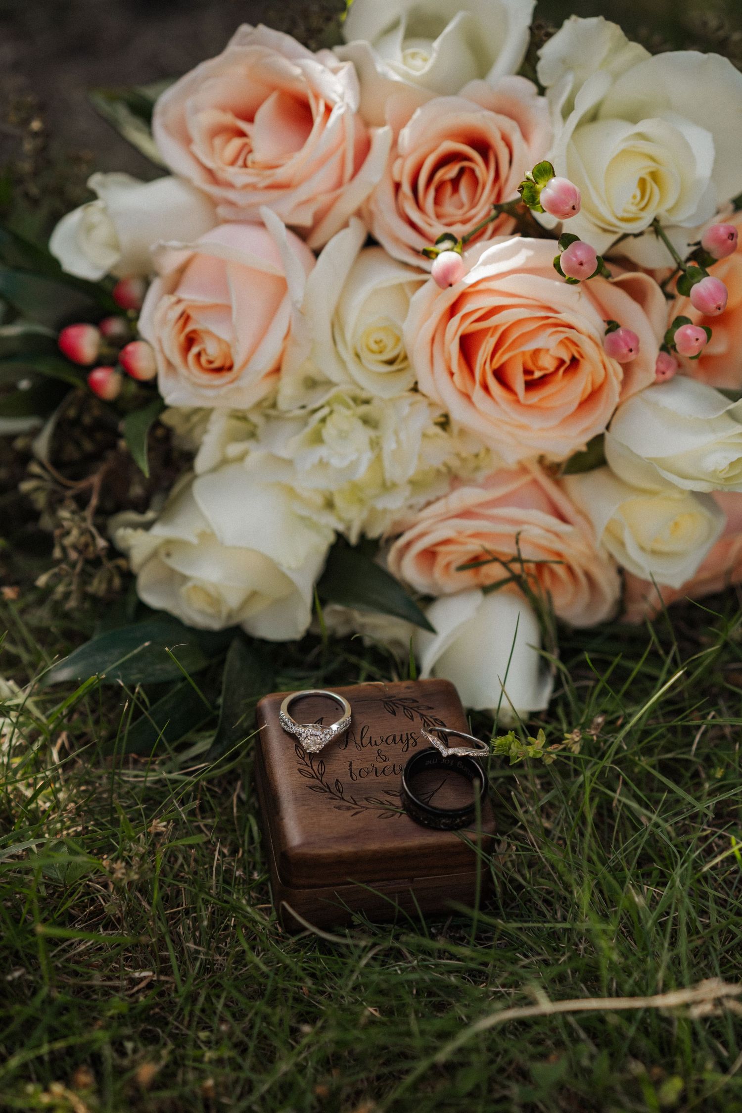 Wedding rings and romantic peach and white roses arranged on grass with wooden accessories.