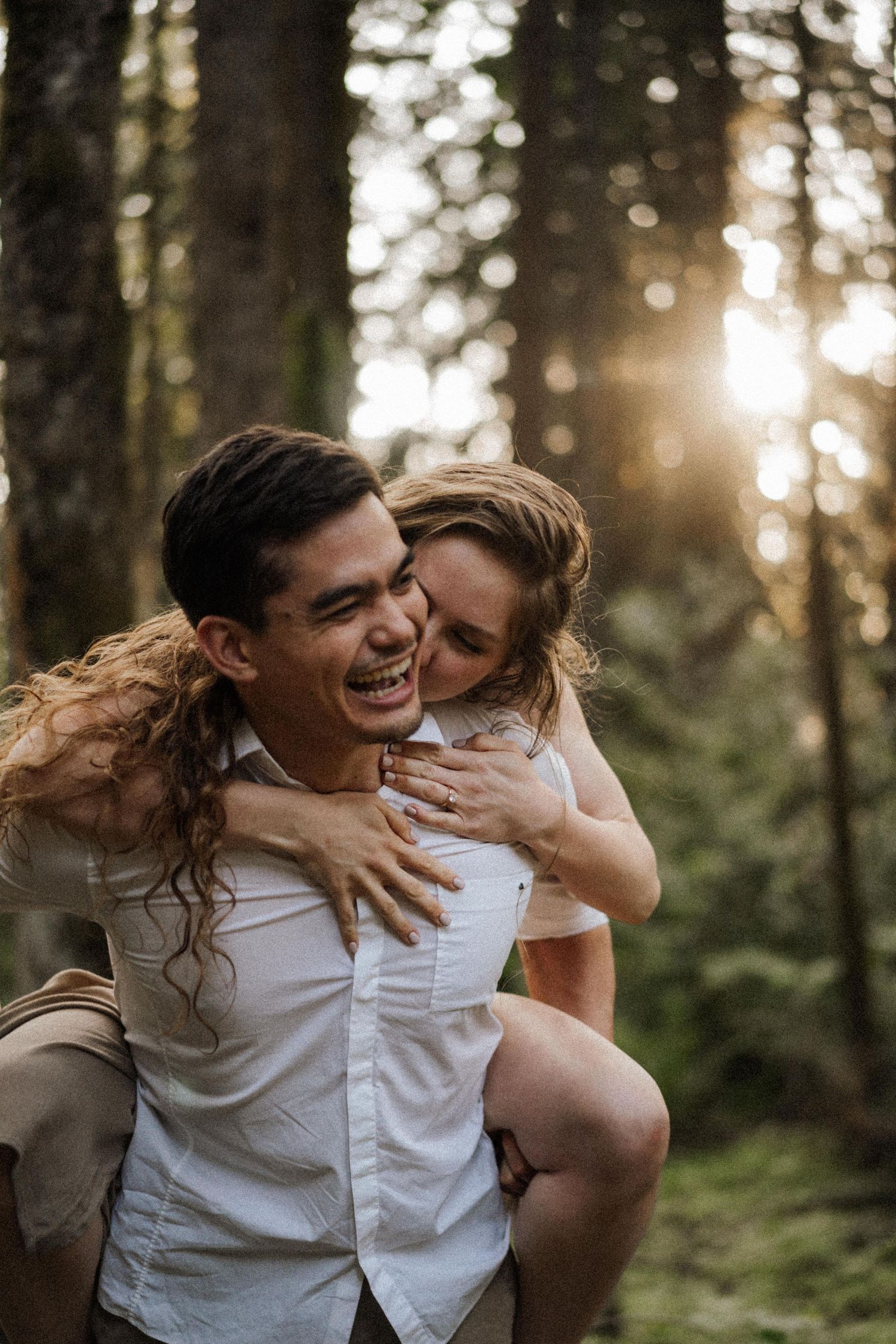 Couple laughing and embracing in a sunlit forest during golden hour.