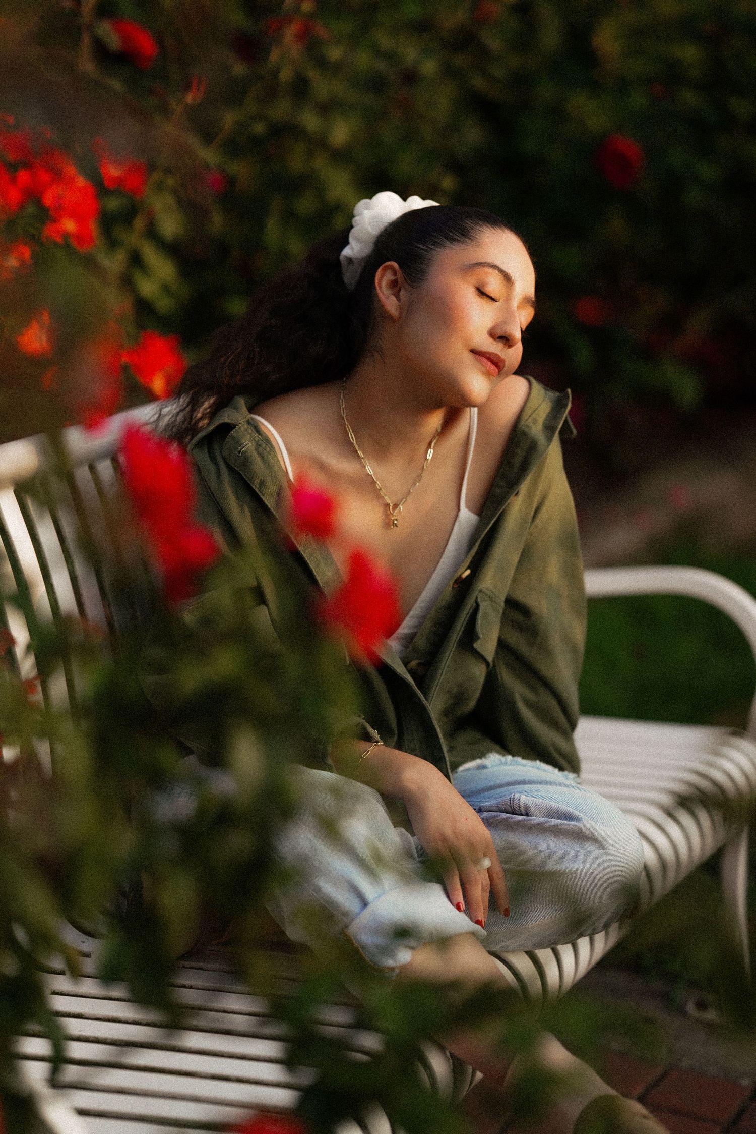 Person in olive jacket relaxes on outdoor bench with red flowers in foreground during golden hour.