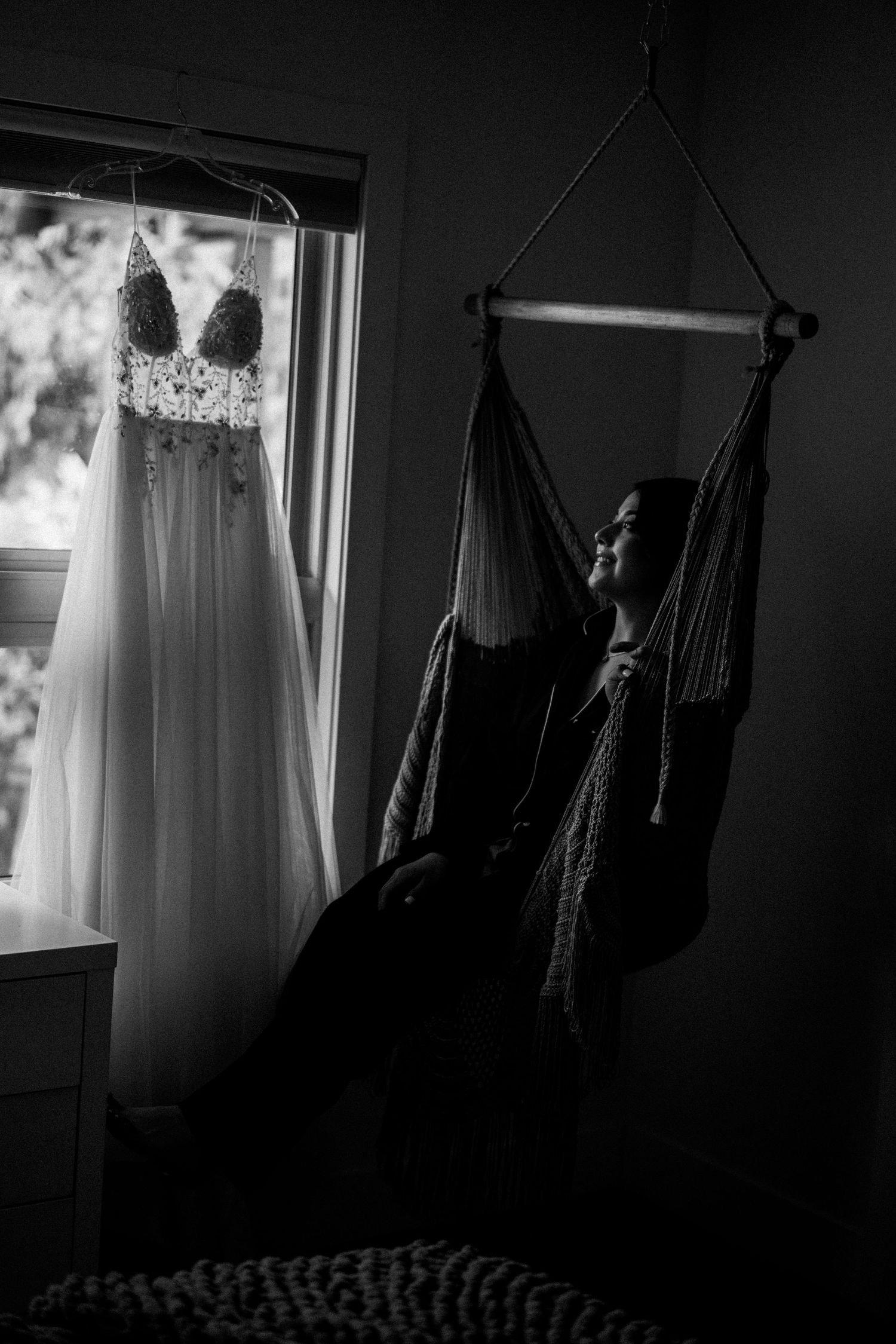 Silhouette of wedding dresses and formal attire hanging by a window in dramatic lighting.