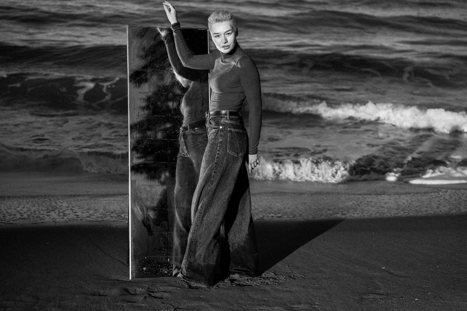 A figure stands on a dark beach holding a staff against a dramatic ocean backdrop in black and white.