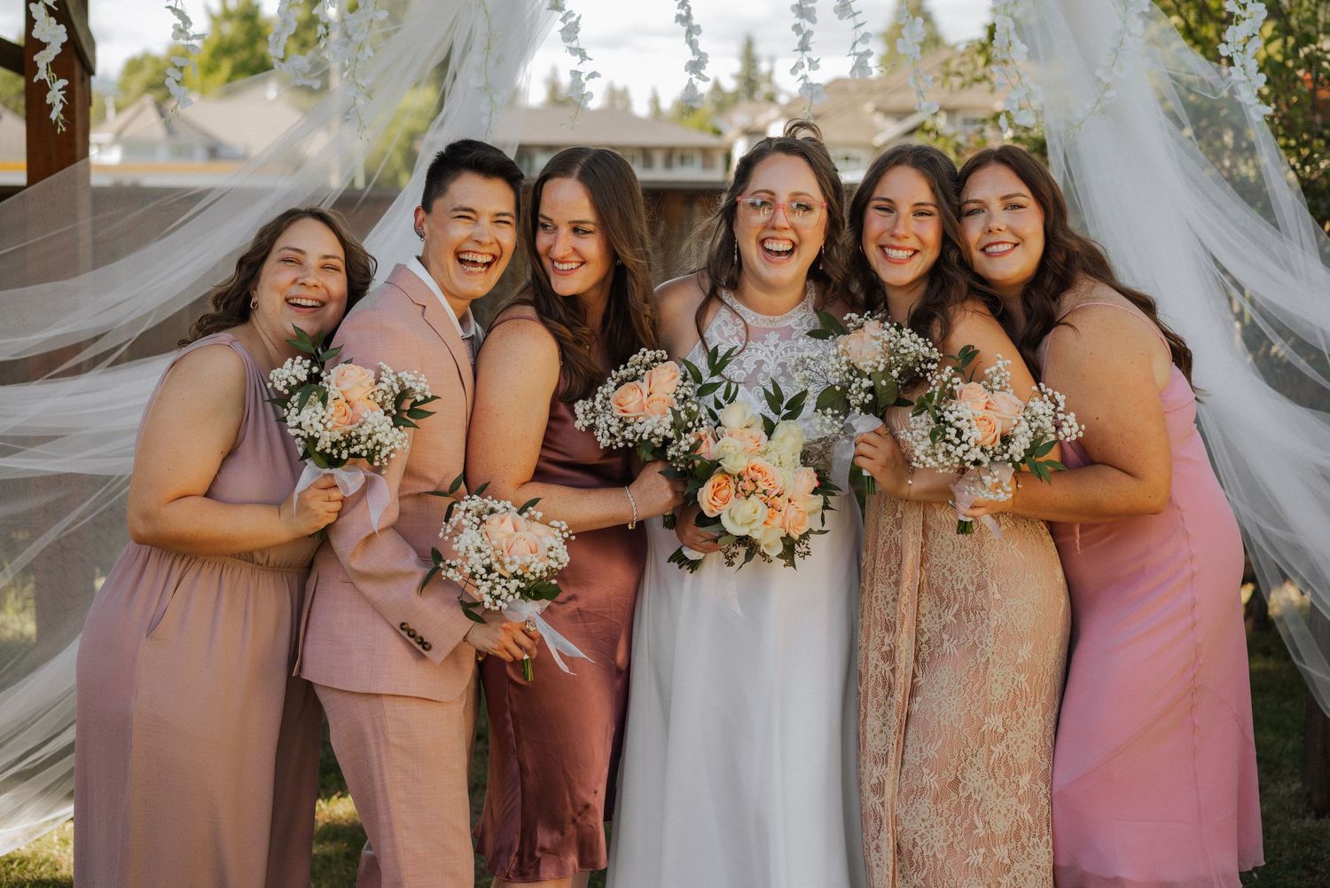 Bridesmaids wearing pink and blush-toned dresses pose together at an outdoor wedding celebration.
