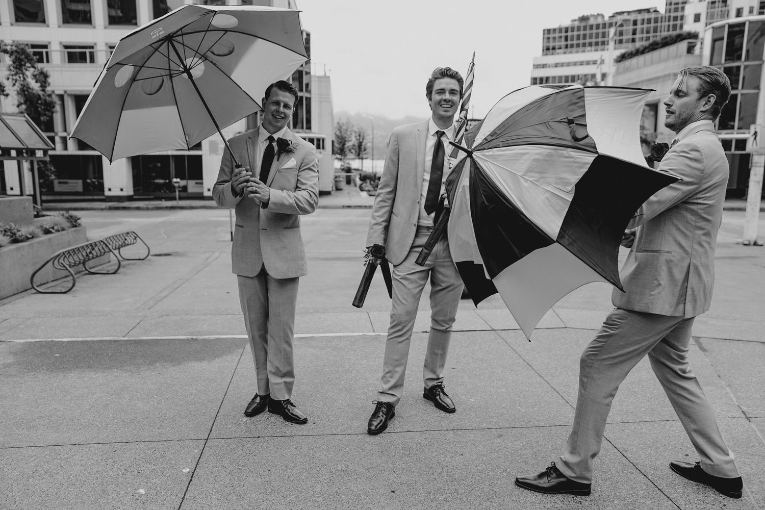 Wedding party walking down city sidewalk holding umbrellas.
