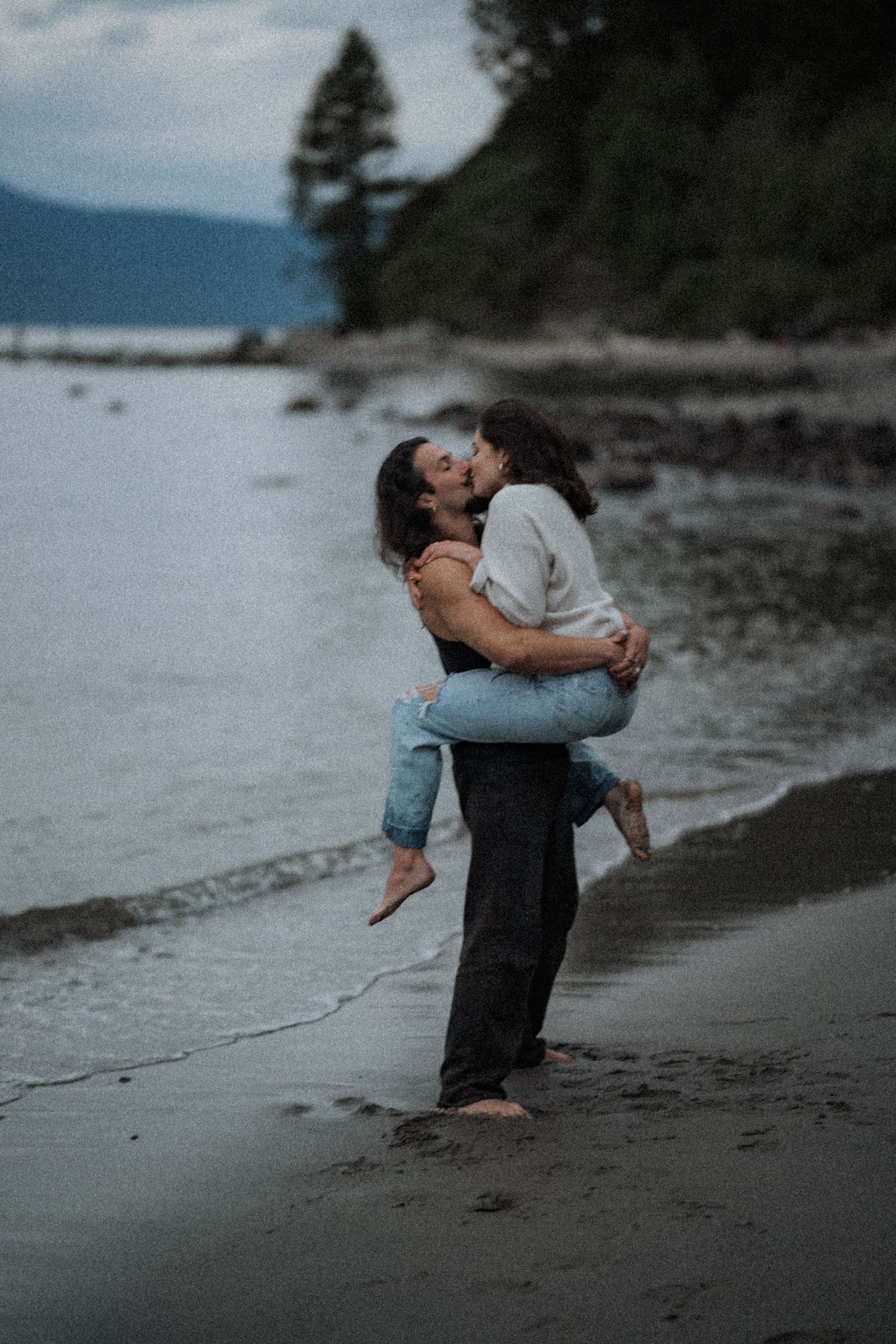 A playful couple embraces on a moody beach with mountains in the background.