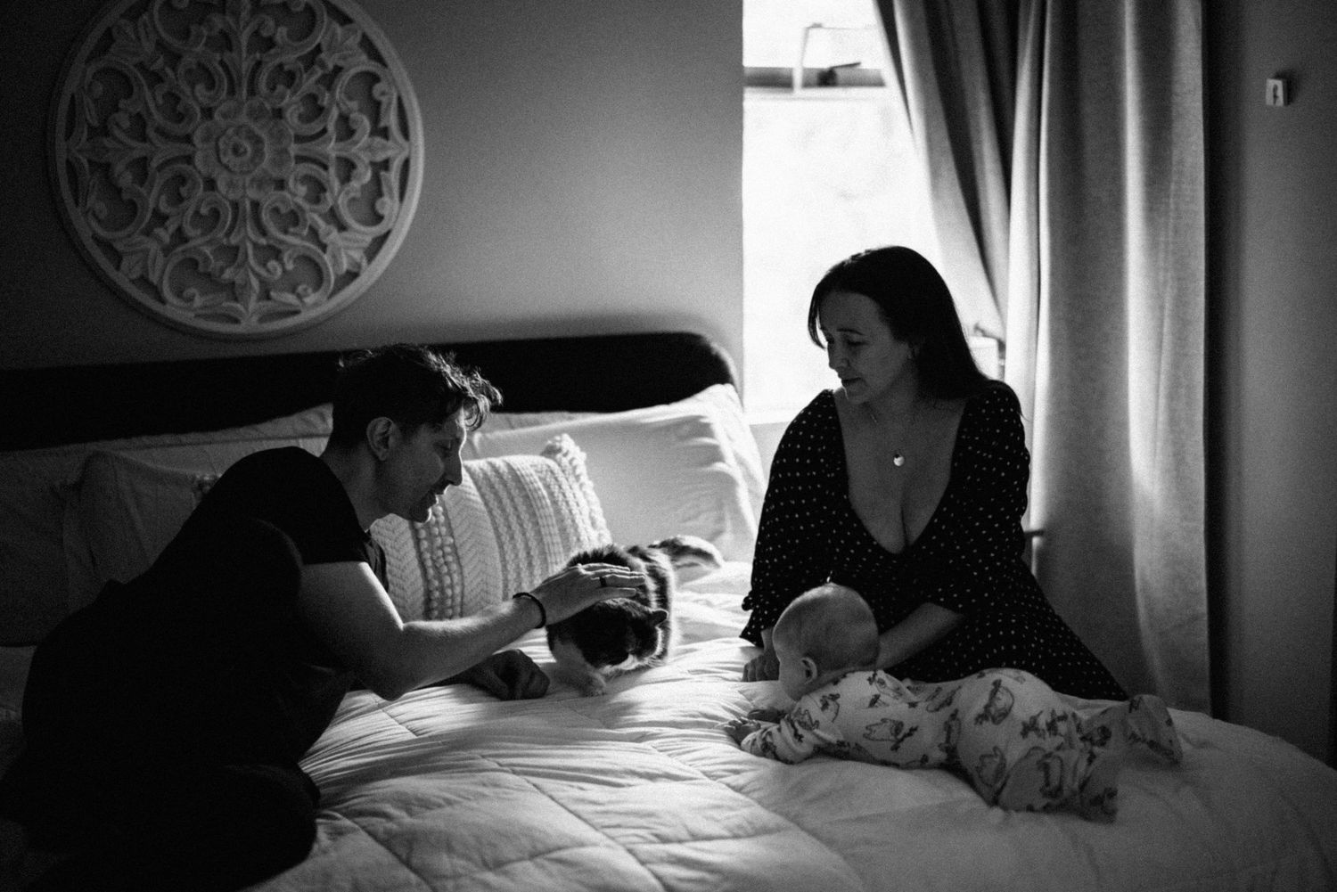 A black and white photograph shows two people sitting on a bed in conversation while a baby sleeps between them.