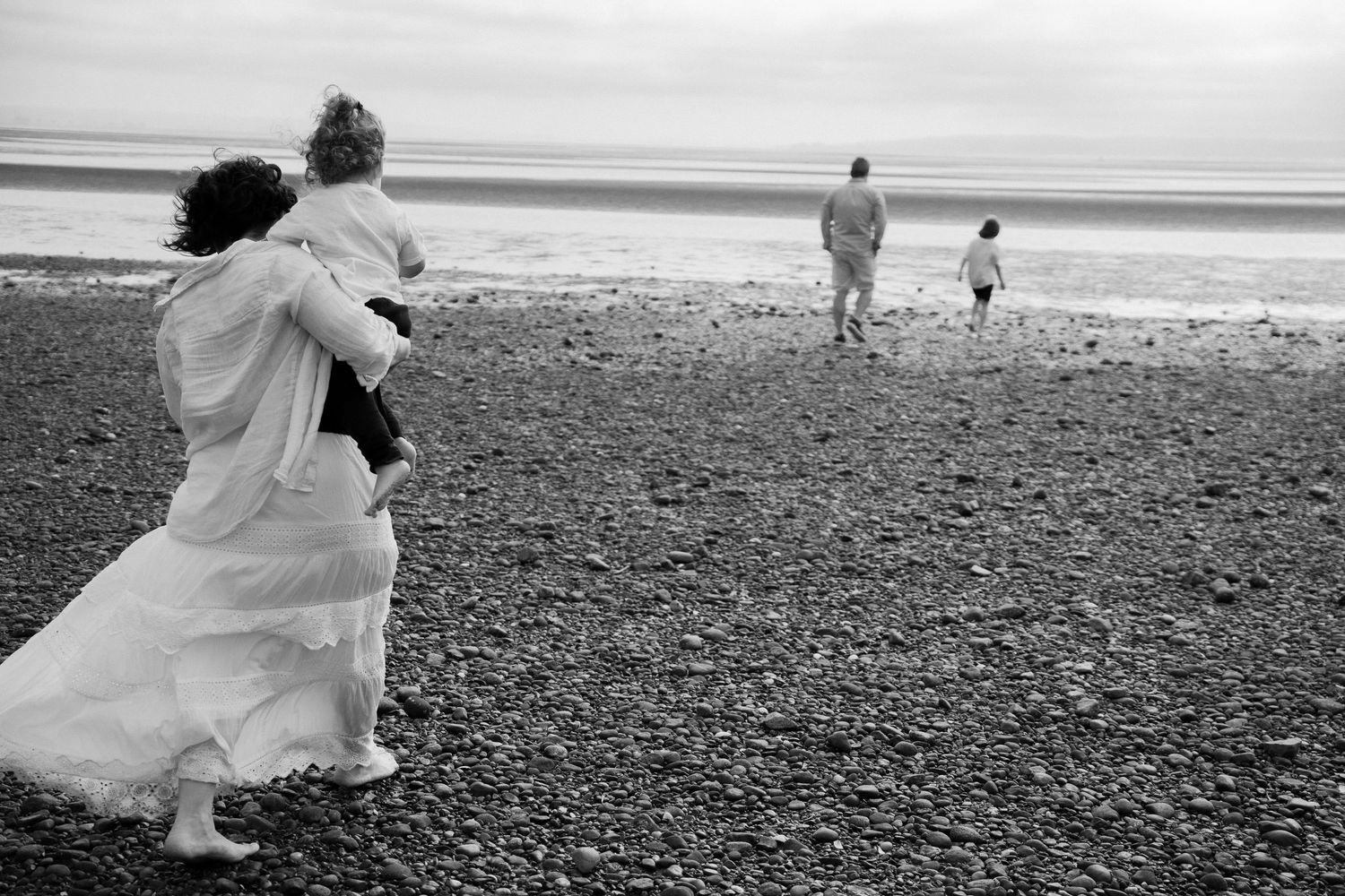 Figures walking along pebbled beach with white dress flowing in the wind.