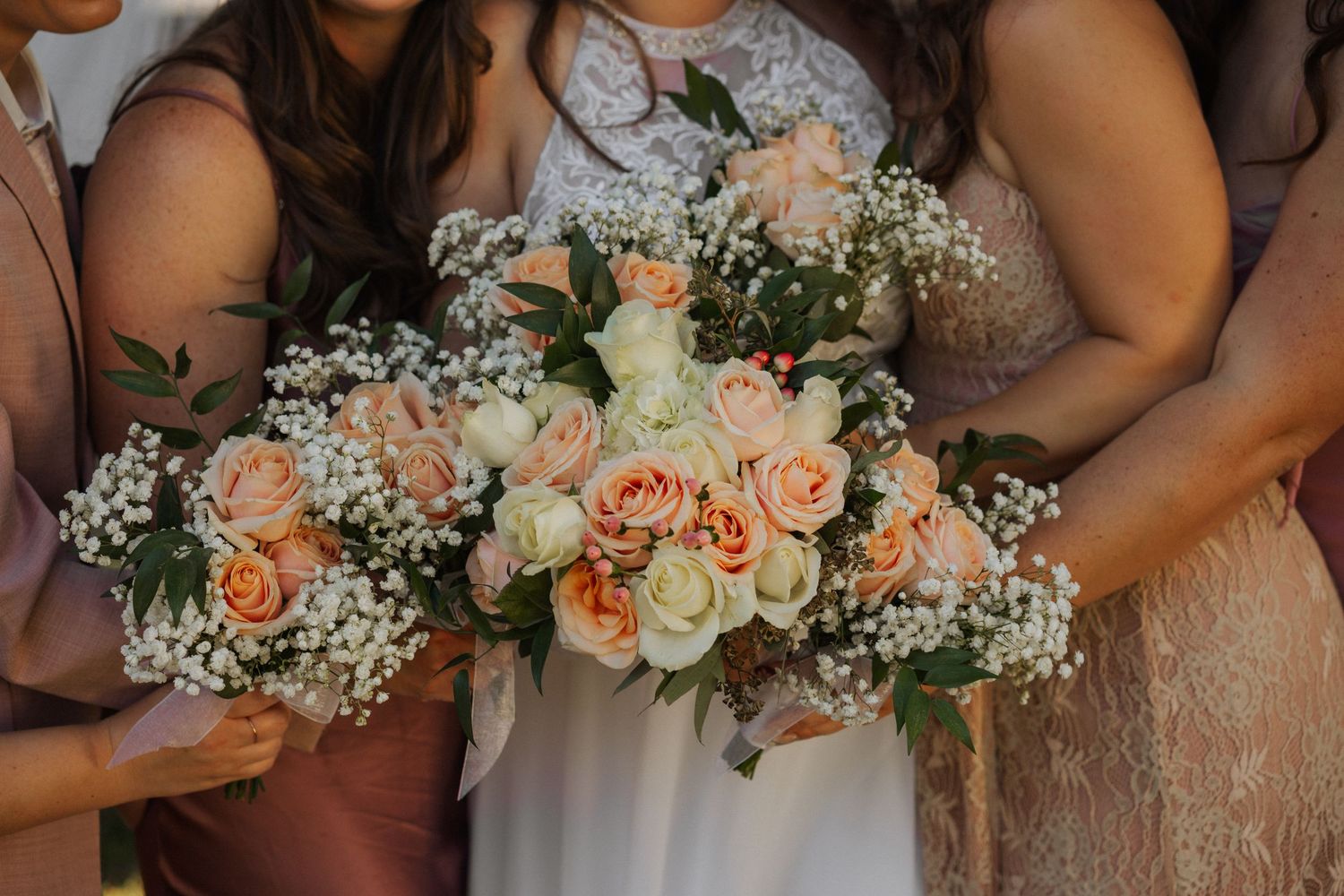 Wedding party members hold elegant peach and ivory floral bouquets with roses and baby's breath against rustic backdrop.