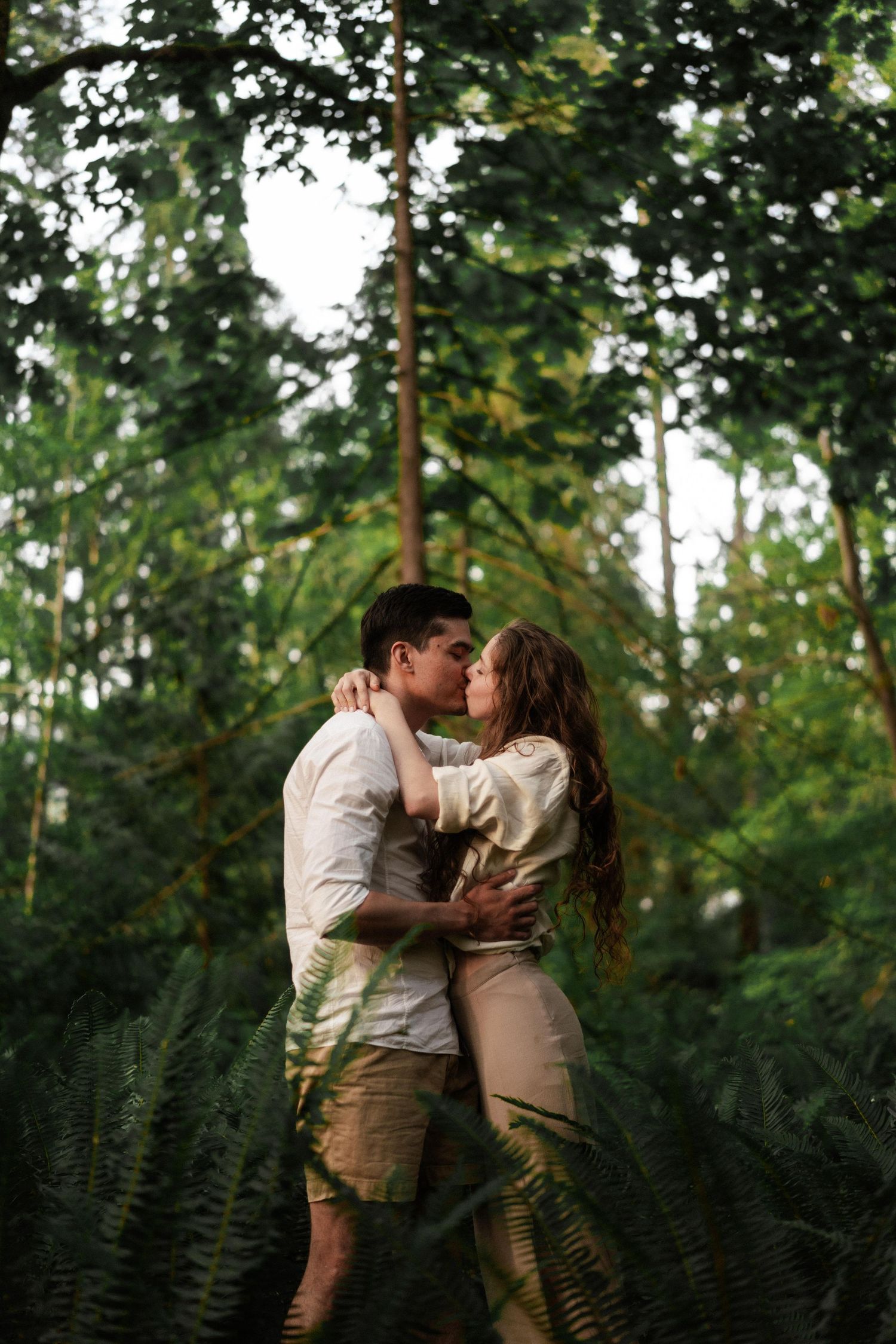 Couple in light colored clothing embraces among ferns in a lush forest setting.