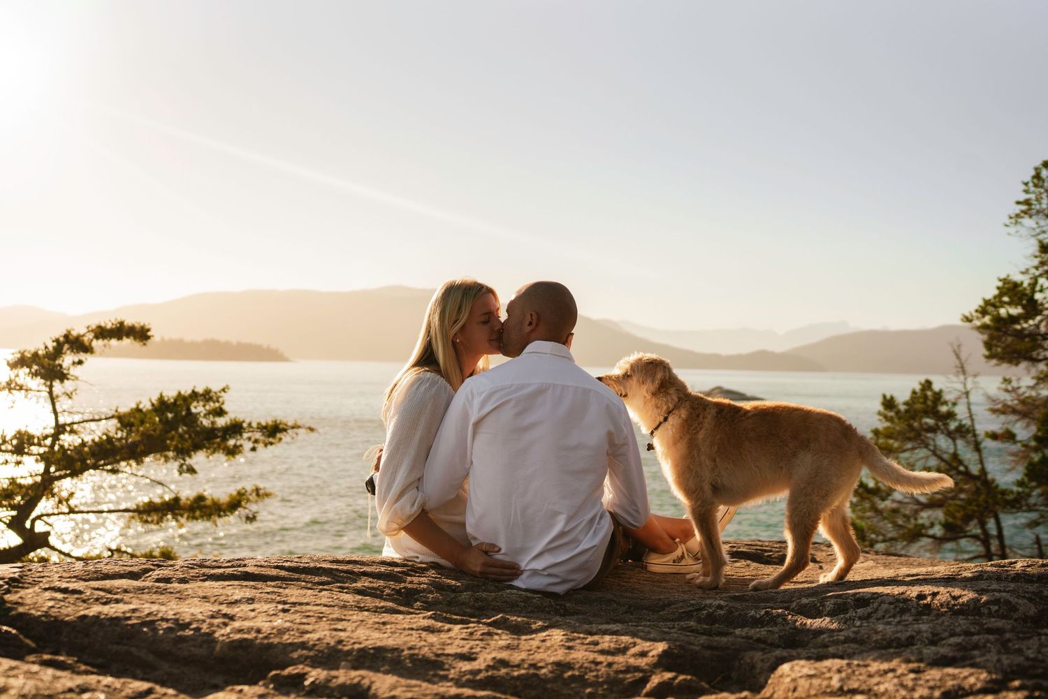 Dog joins couple watching sunset from rocky coastal viewpoint.