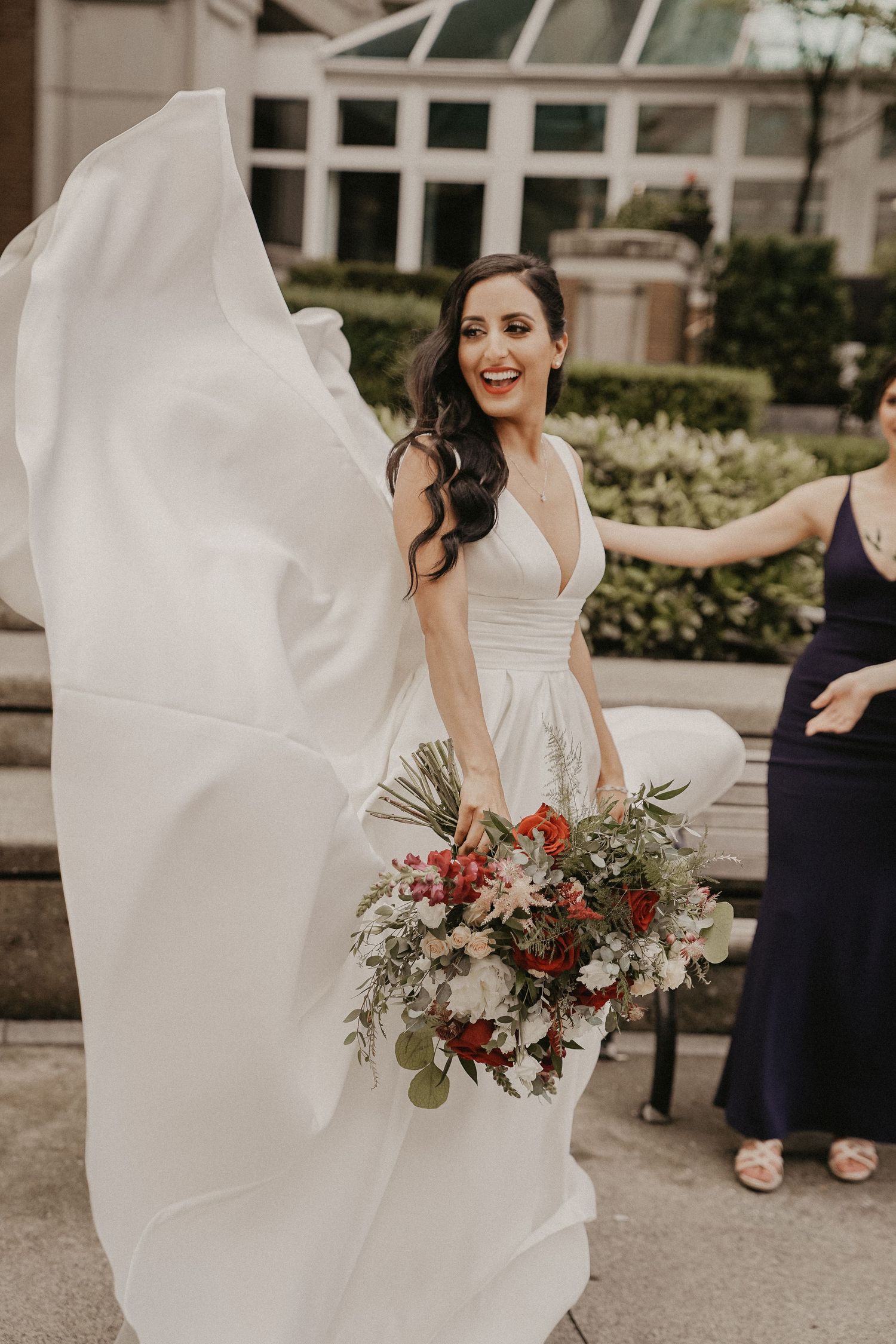 A bride in a flowing white dress holds a burgundy and green bouquet while her veil catches the breeze outdoors.