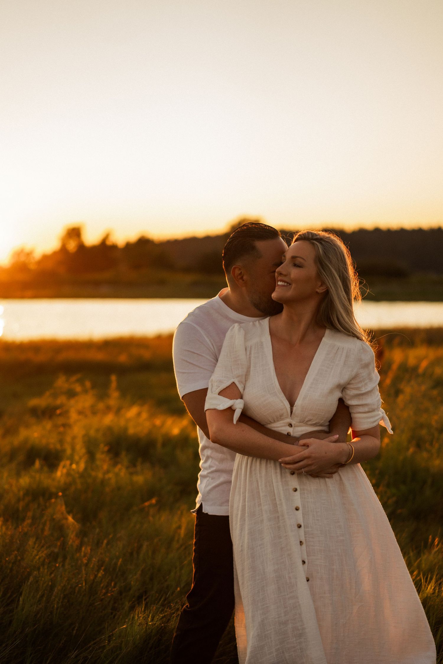 Couple shares intimate moment in golden sunlit field by water.