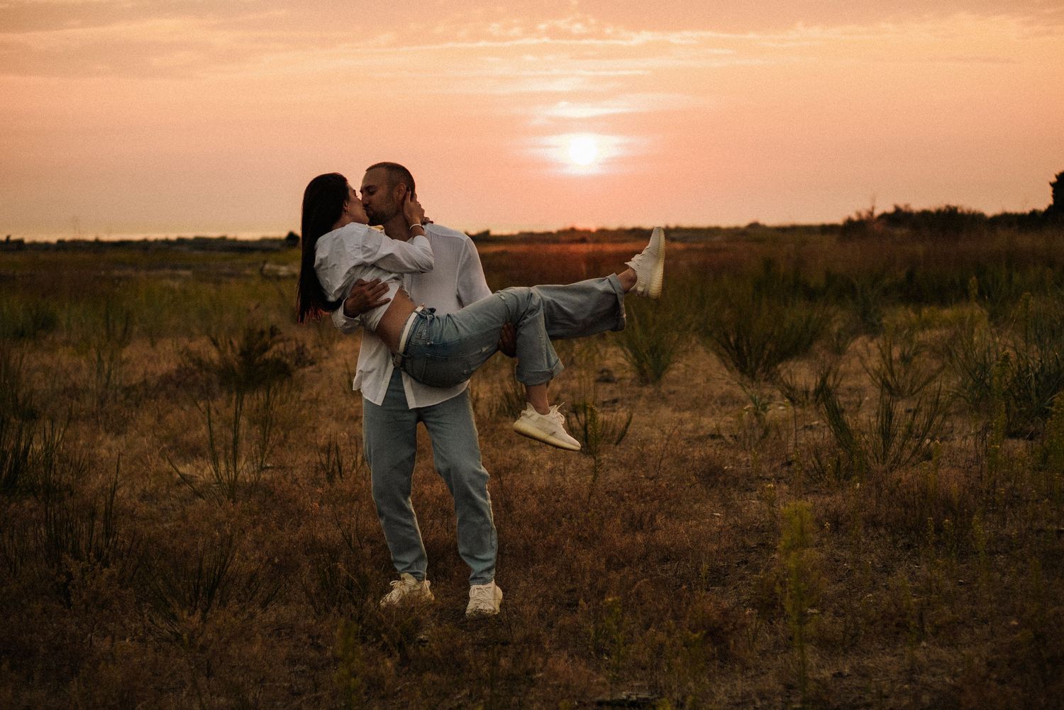 A couple shares a romantic moment in a field at sunset as one person carries the other.