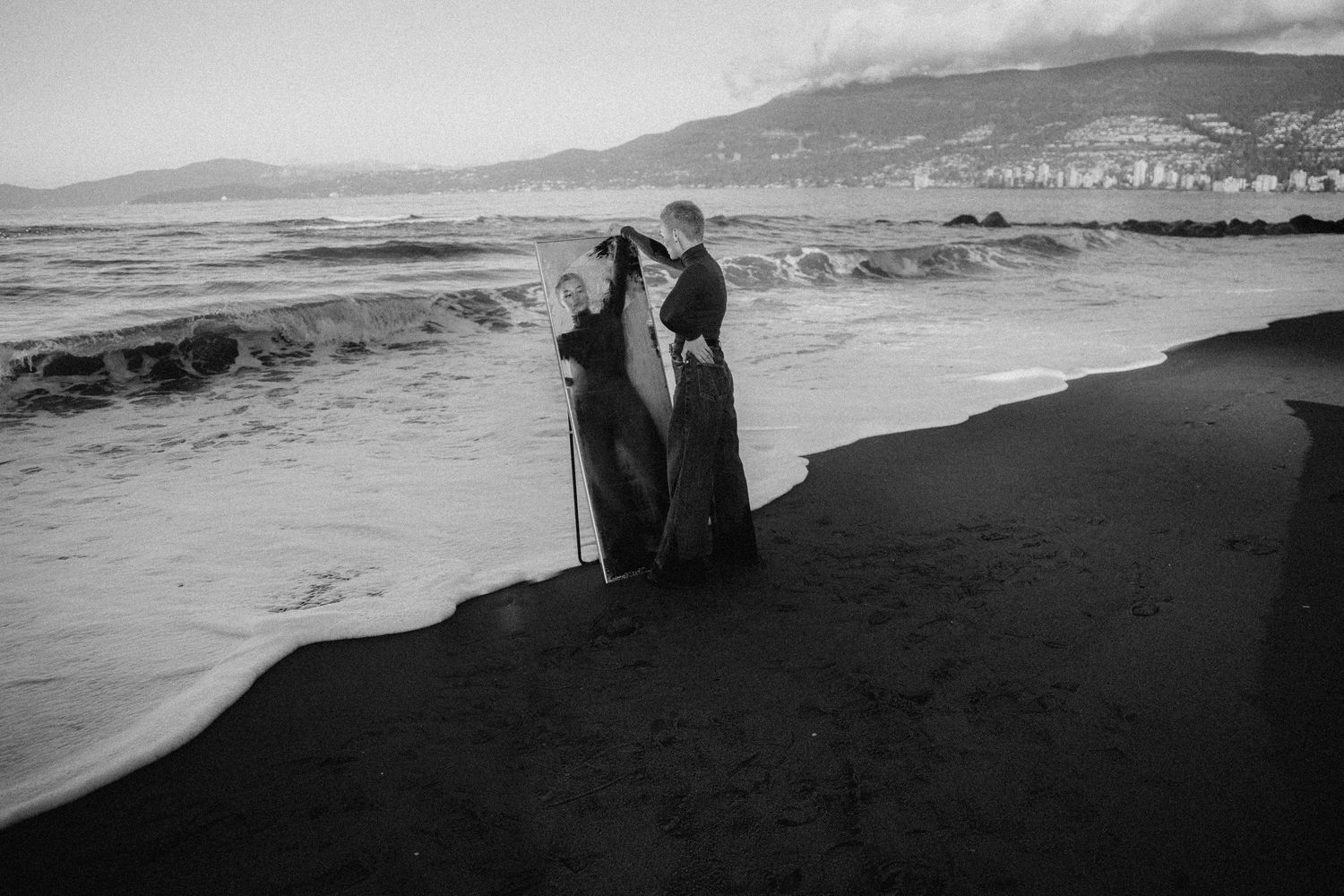 Two silhouettes stand together on a black sand beach with waves and mountains in the distance, captured in black and white.