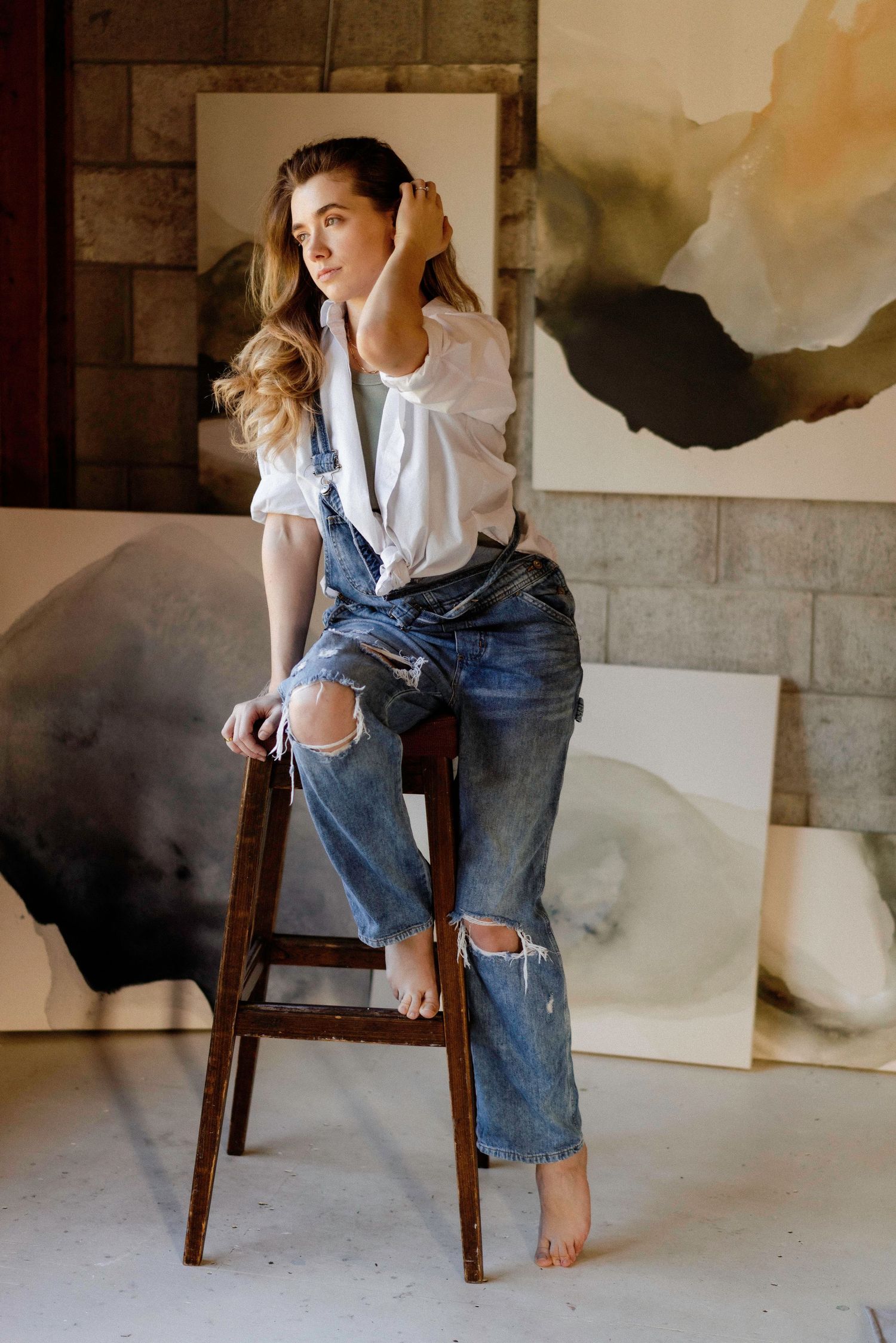 Person in distressed jeans and white shirt sitting casually on wooden chair in artistic setting.