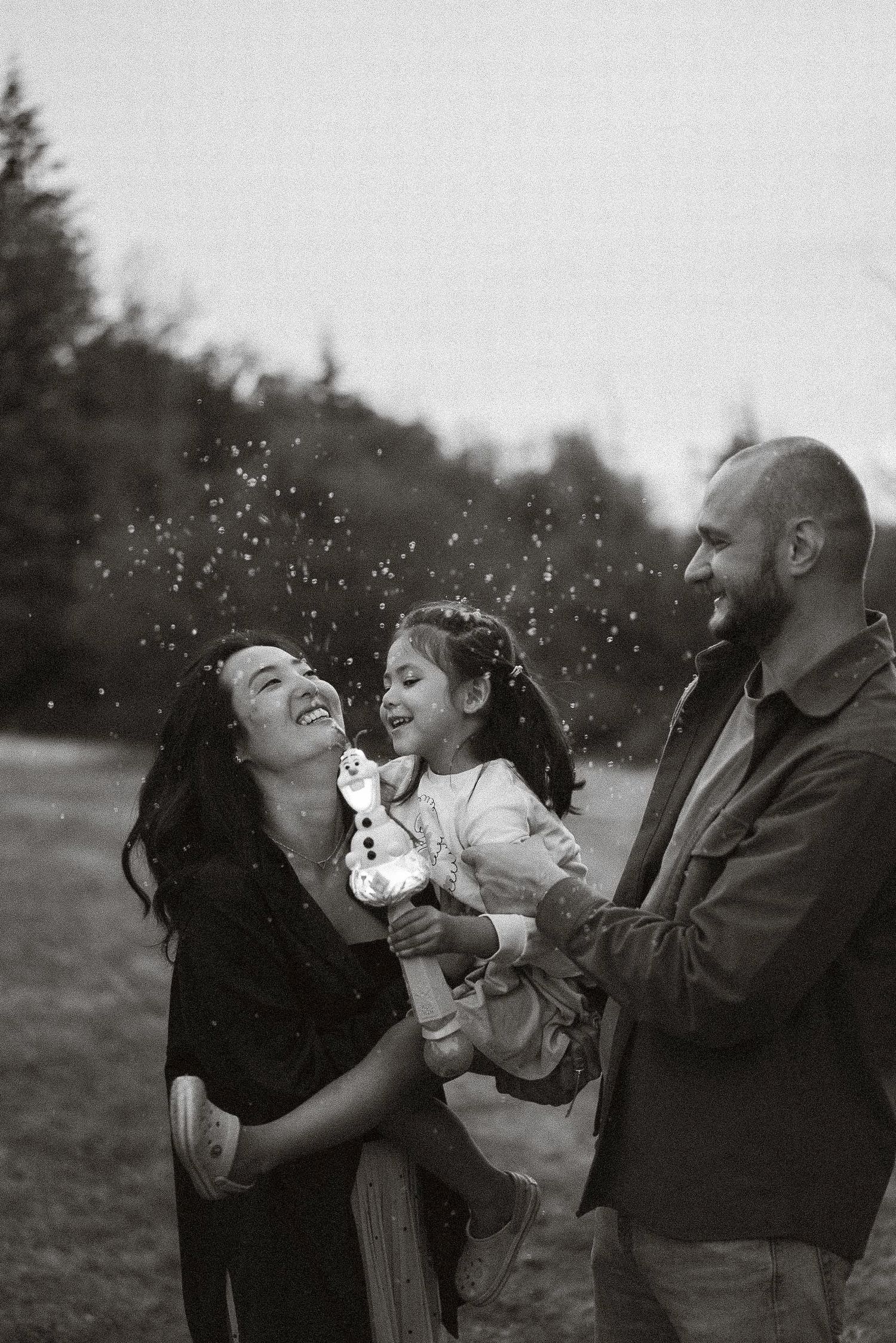 A candid black and white photo sequence shows a family playing and laughing together outdoors in nature.