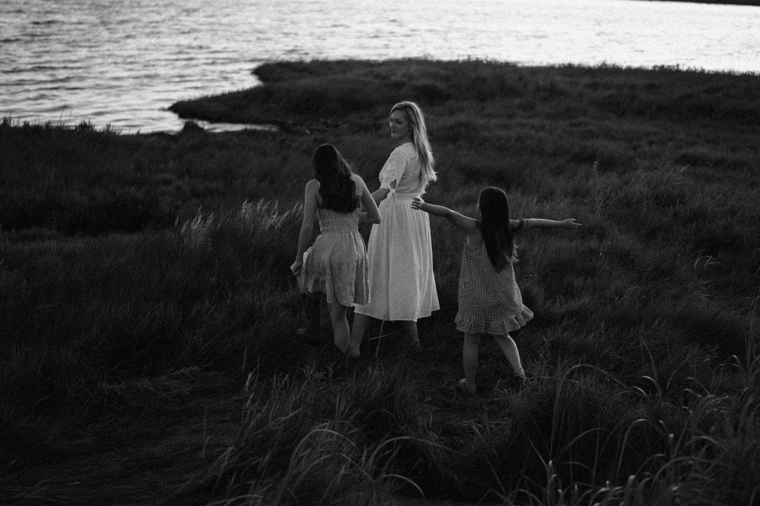 Three silhouettes stand along grassy shoreline during twilight in black and white photography.