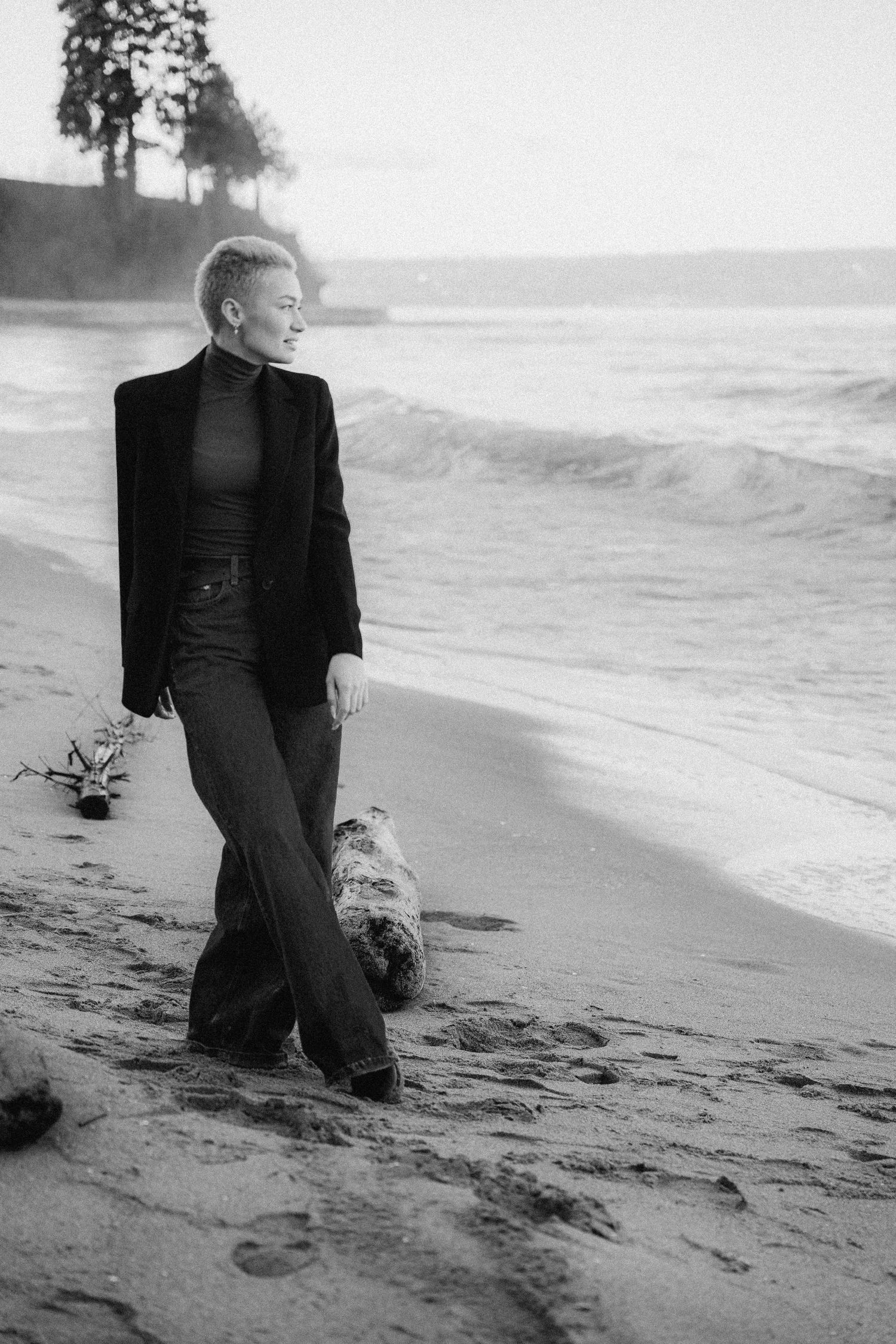 Black and white portrait of a person in dark clothing standing thoughtfully on a beach at sunset.
