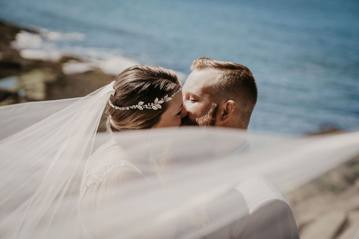 Bride's white veil flows in the wind as couple shares romantic moment near ocean coastline.