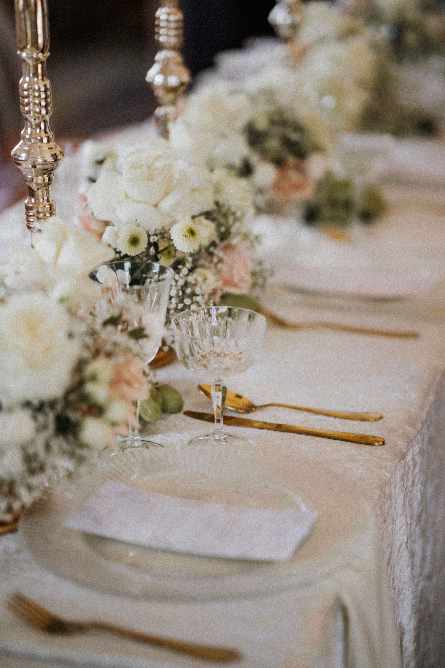 Elegant wedding reception table setting with delicate white floral centerpieces, crystal glassware and gold utensils.