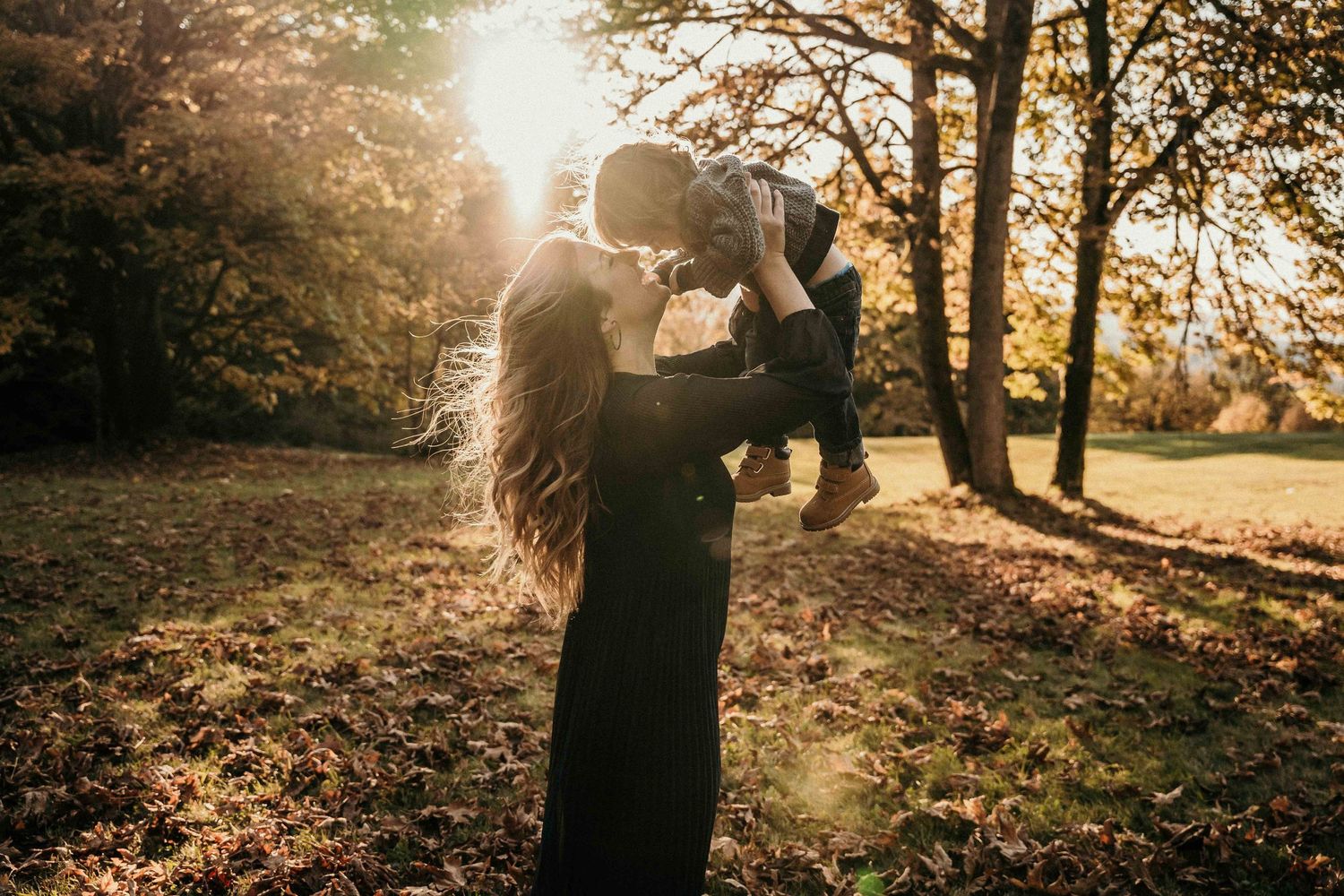 A silhouette holds up a small child against golden sunlight streaming through autumn trees in a park.