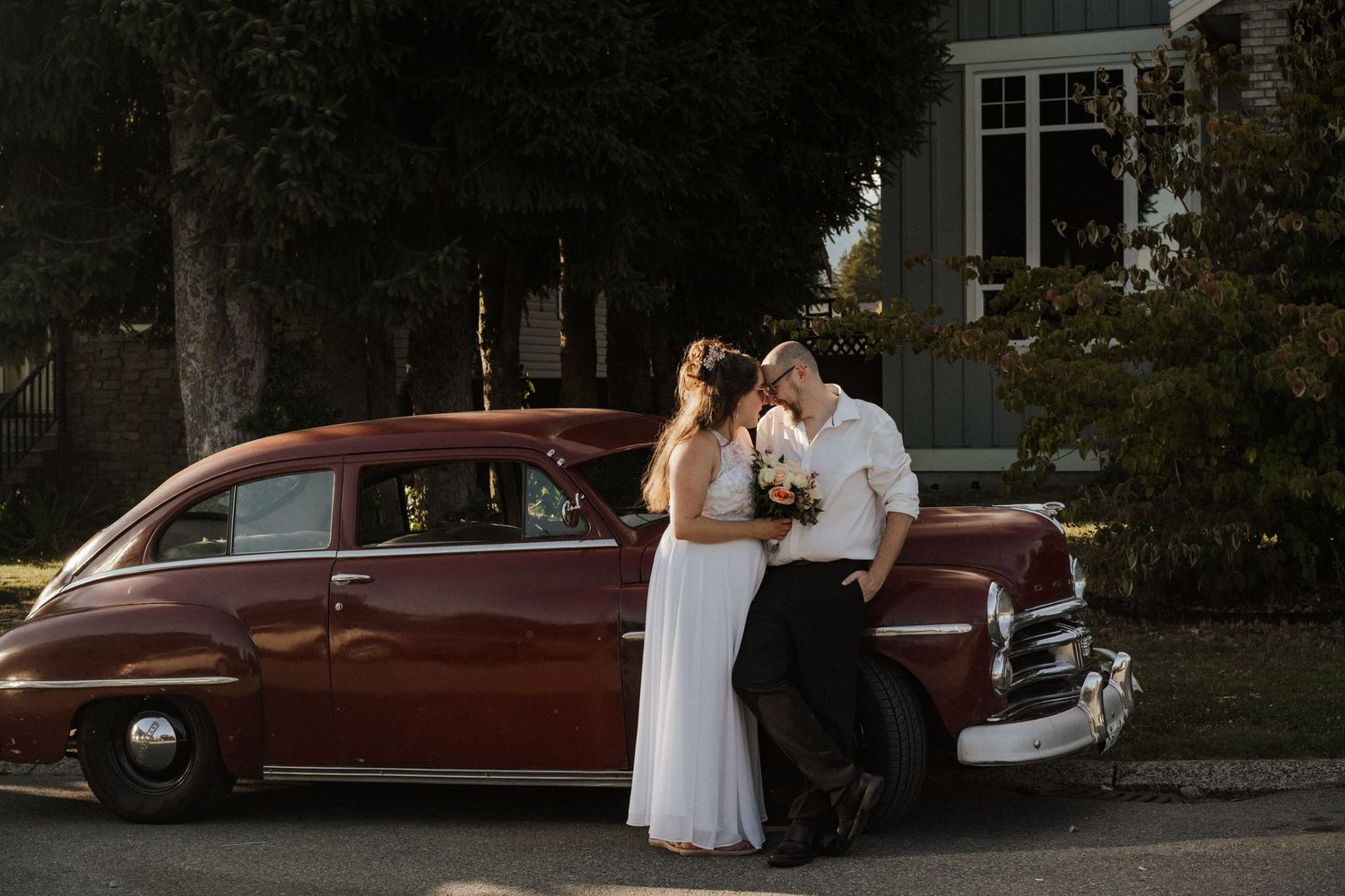 A couple in wedding attire poses near a vintage burgundy car during their outdoor photo session.